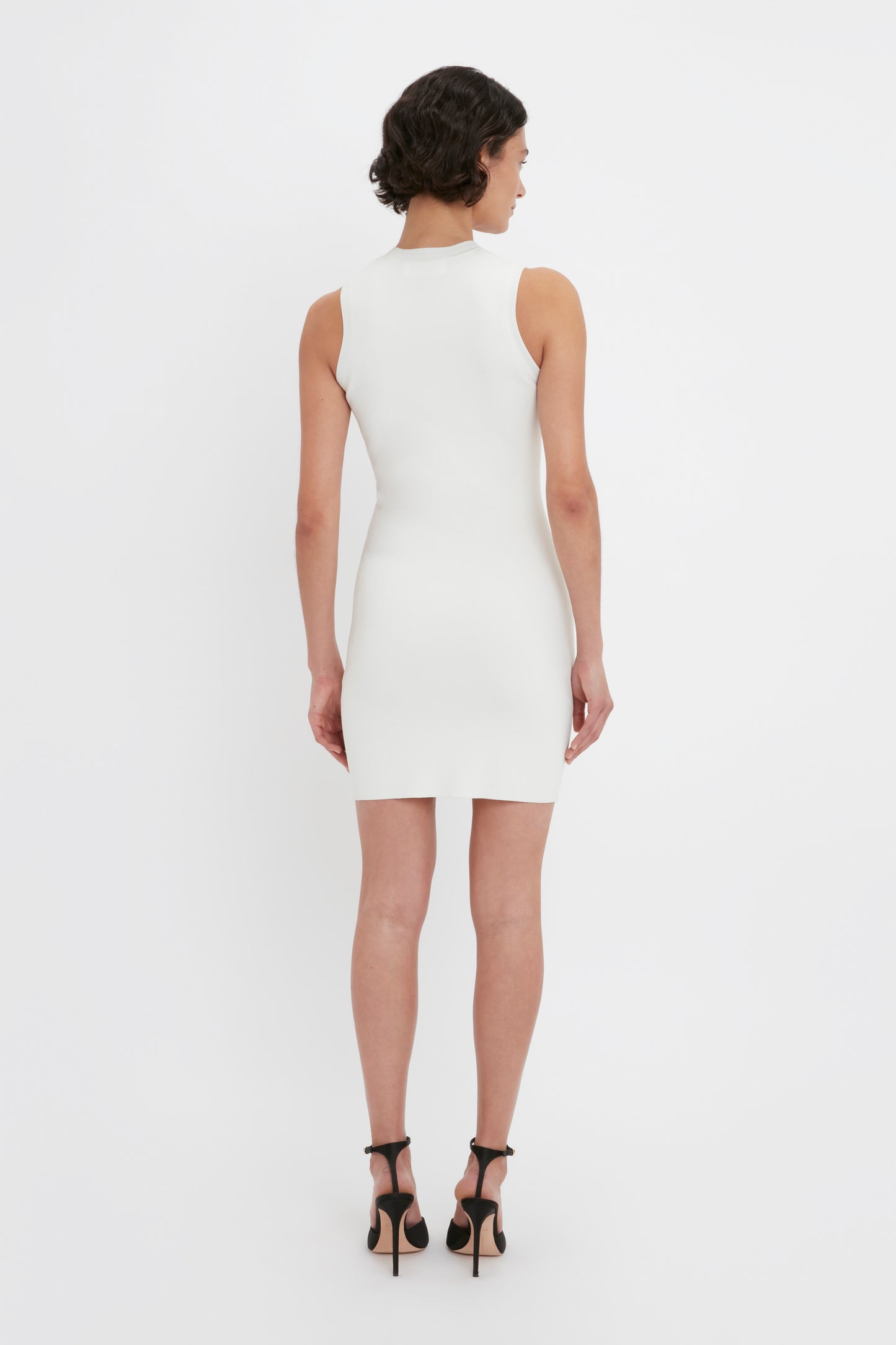 A person is standing facing away from the camera, wearing the VB Body Mini Dress in White from Victoria Beckham and black high heels, set against a plain white background.