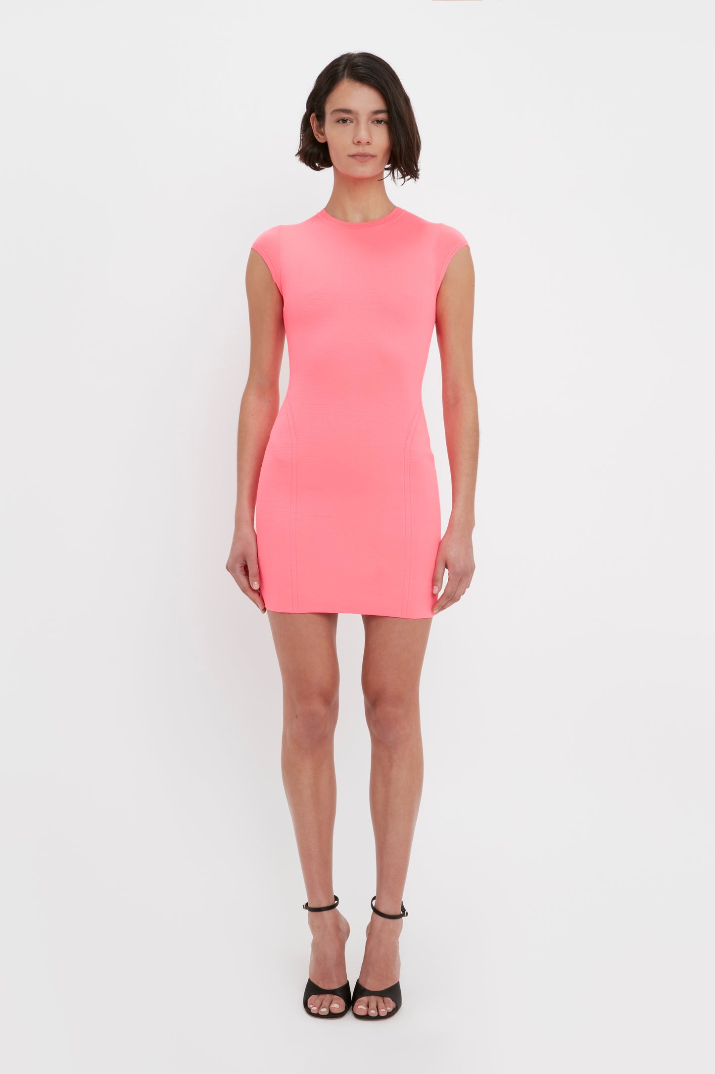 A person stands against a white background wearing the Victoria Beckham VB Body Compact Cap Sleeve Mini Dress in Pink and black high-heeled sandals.