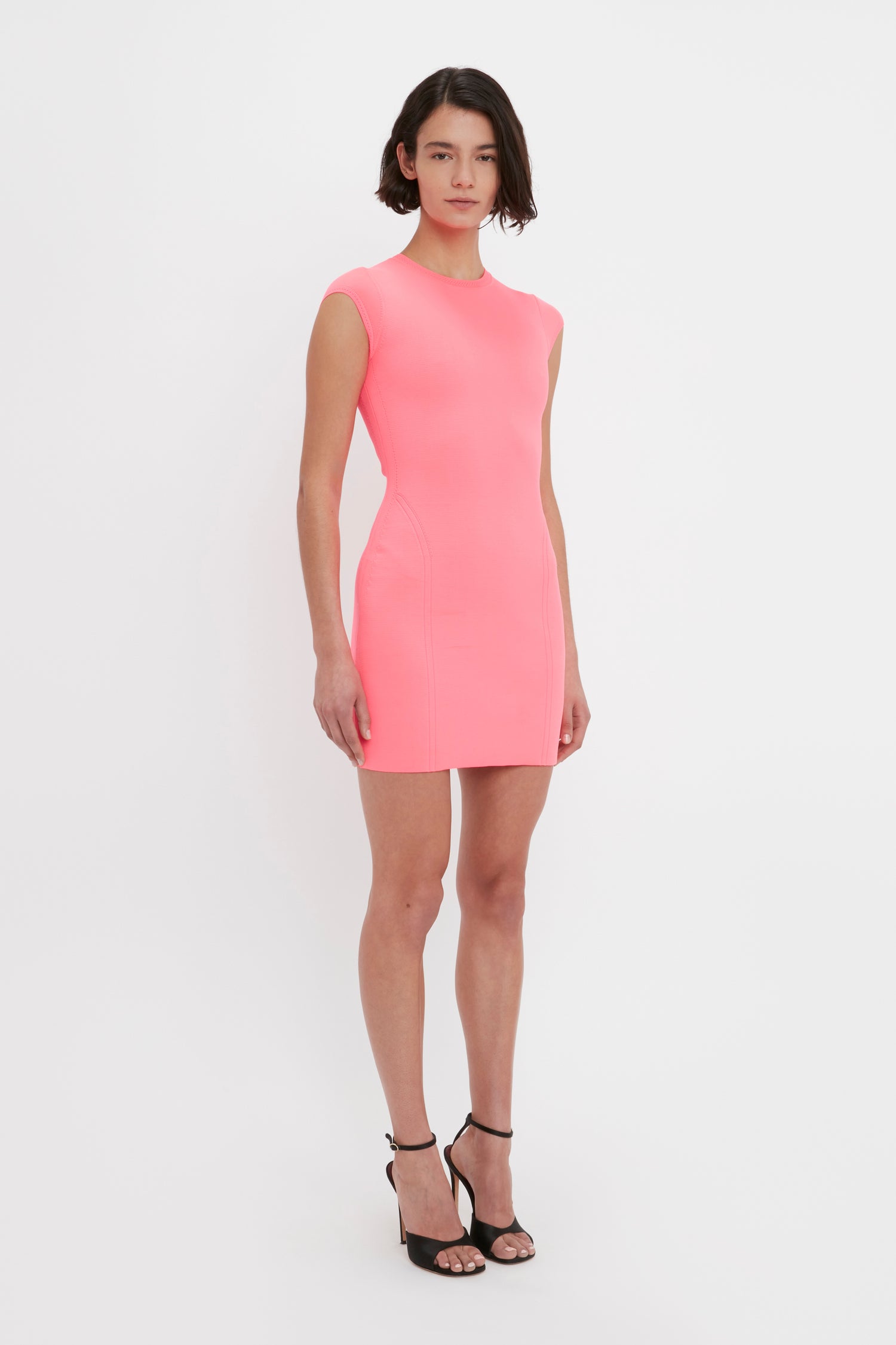 A person with short dark hair is wearing a VB Body Compact Cap Sleeve Mini Dress in Pink by Victoria Beckham and black high-heeled sandals, standing against a plain white background.