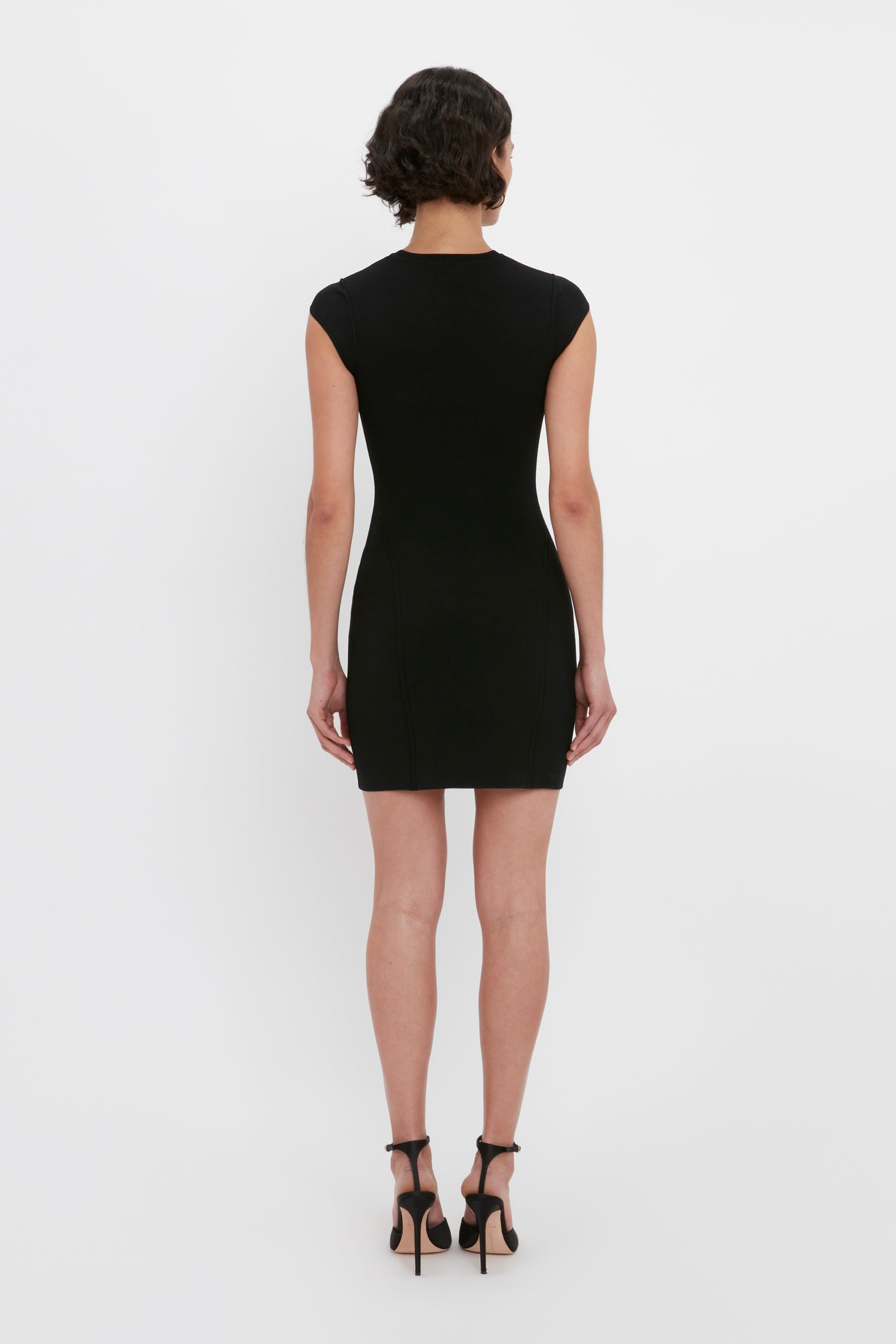 A person with short, curly hair is standing with their back to the camera, wearing the Victoria Beckham VB Body Compact Cap Sleeve Mini Dress in Black and black high heels.
