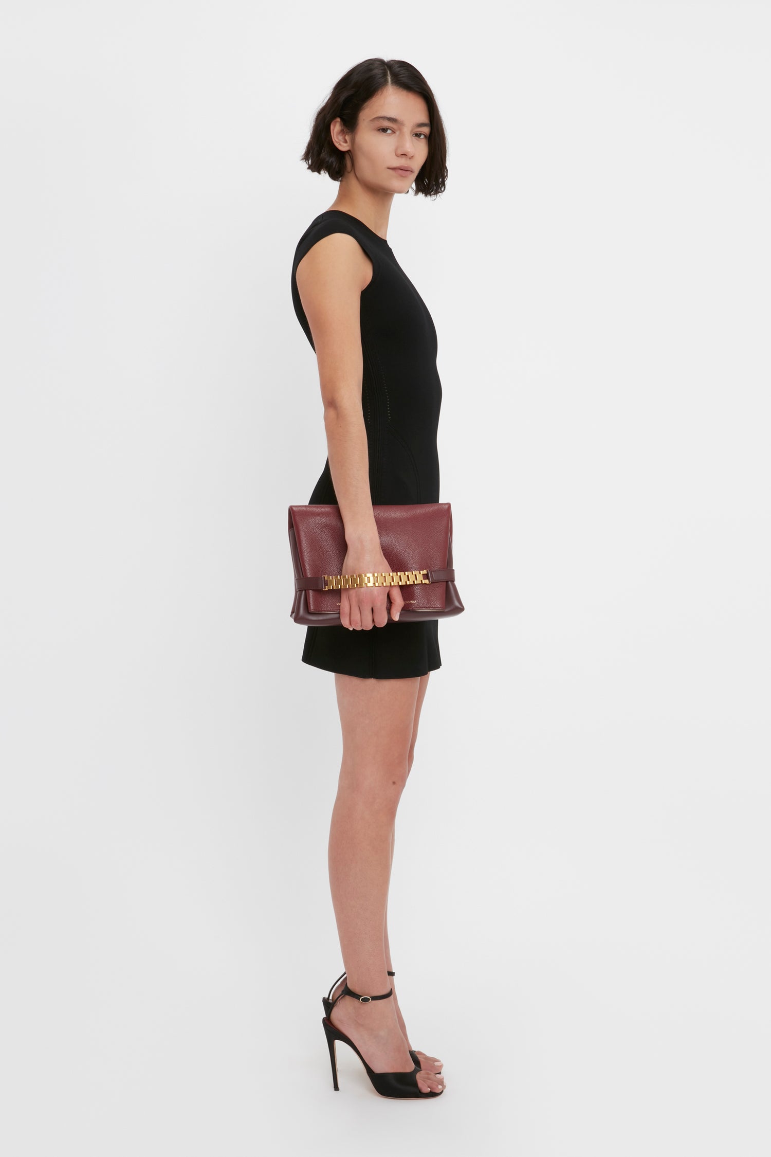 A woman in a VB Body Compact Cap Sleeve Mini Dress in Black by Victoria Beckham and black high heels holds a maroon clutch. She stands against a plain white background.