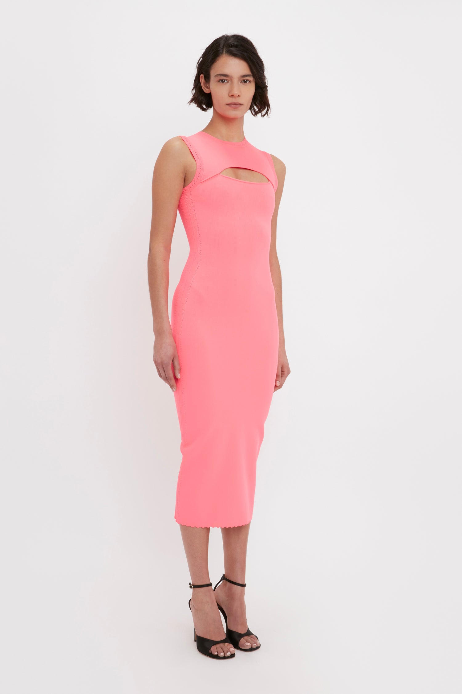 Person standing against a plain white background, wearing the VB Body Slash Front Midi Dress in Pink from Victoria Beckham. The dress is sleeveless, knee-length with a high neckline and features a stylish cutout across the chest. They are also wearing high-heeled sandals.