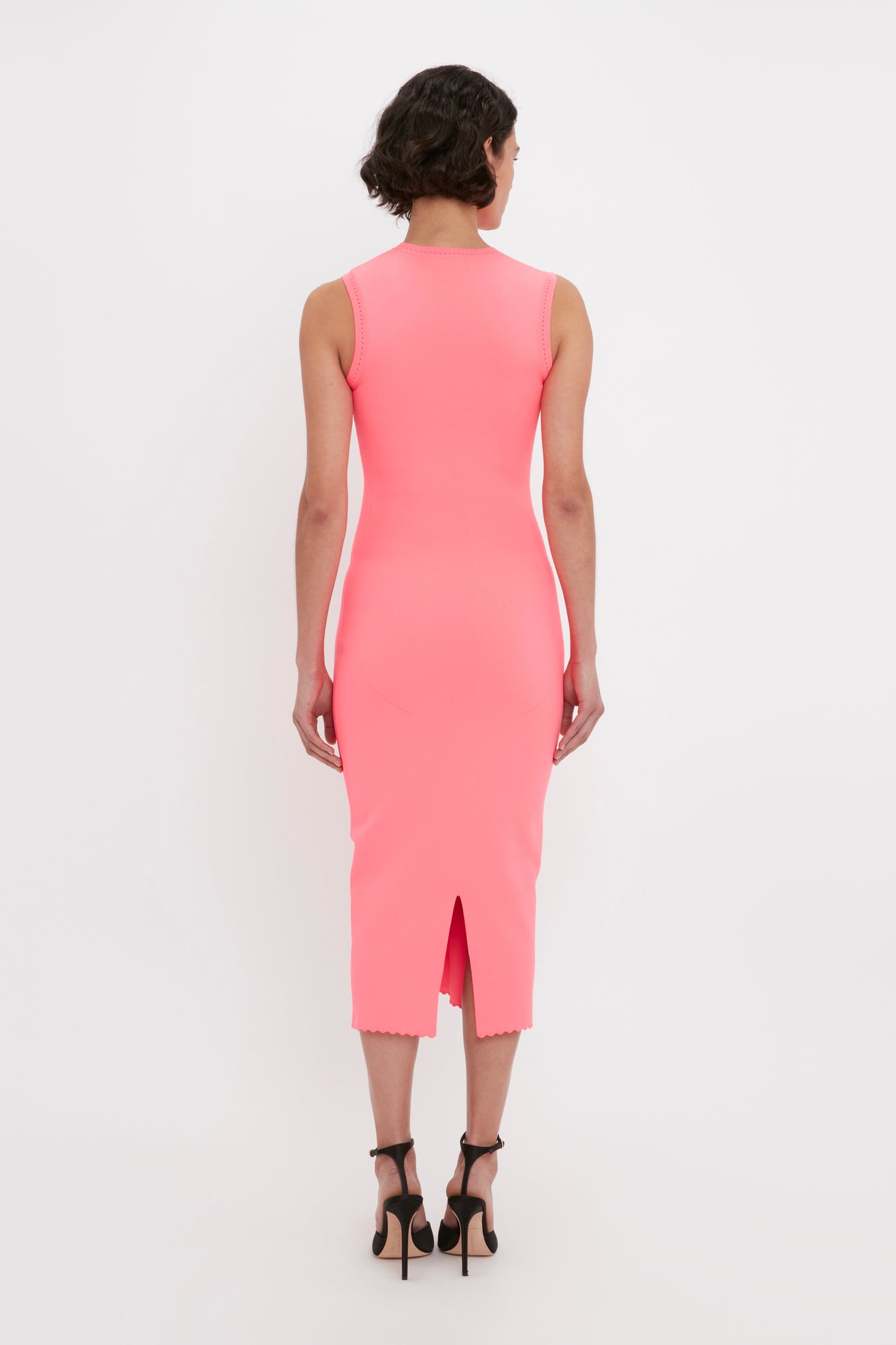 A person with short hair stands facing away, wearing the Victoria Beckham VB Body Slash Front Midi Dress in Pink and black high-heeled shoes.