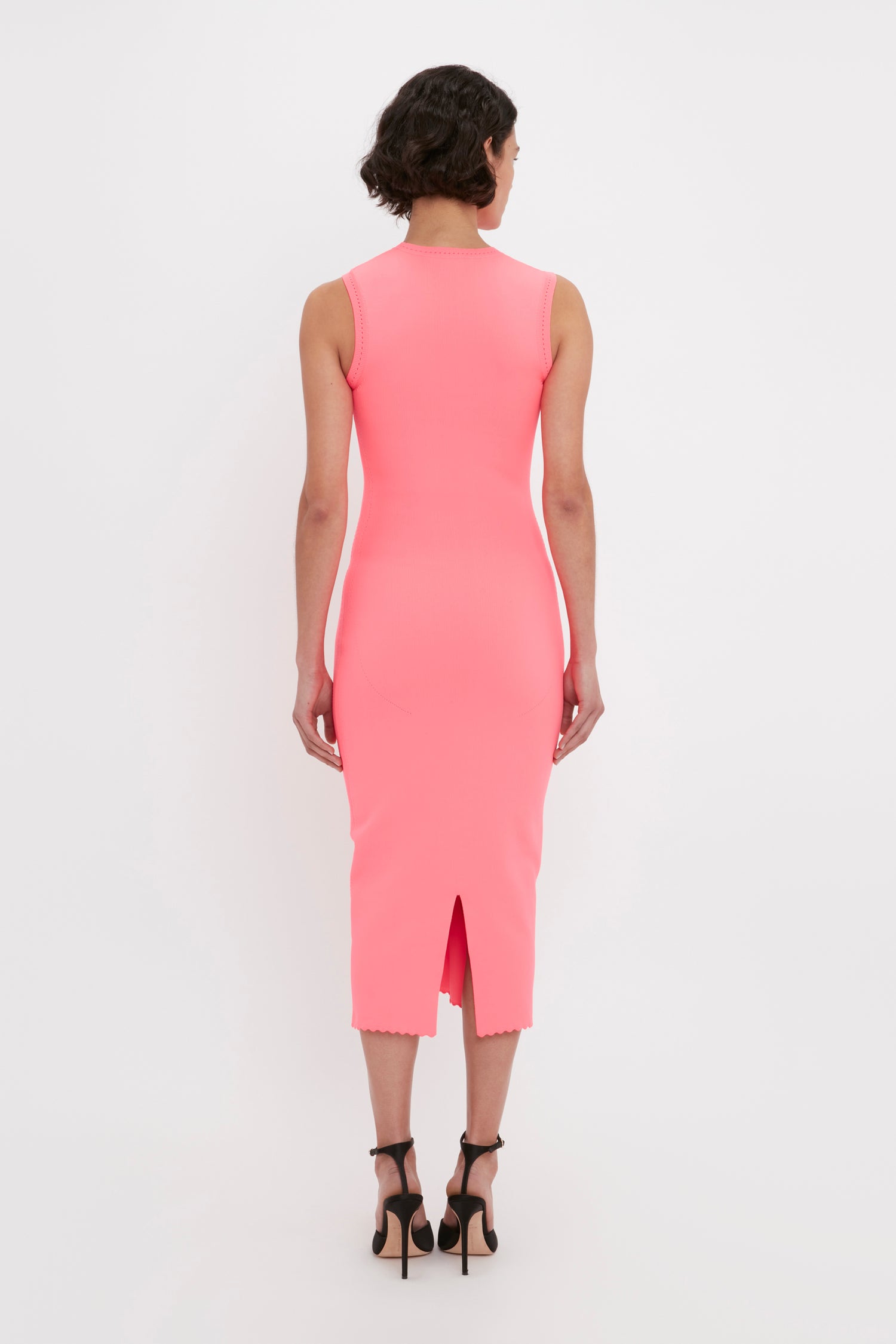 A person with short hair stands facing away, wearing the Victoria Beckham VB Body Slash Front Midi Dress in Pink and black high-heeled shoes.