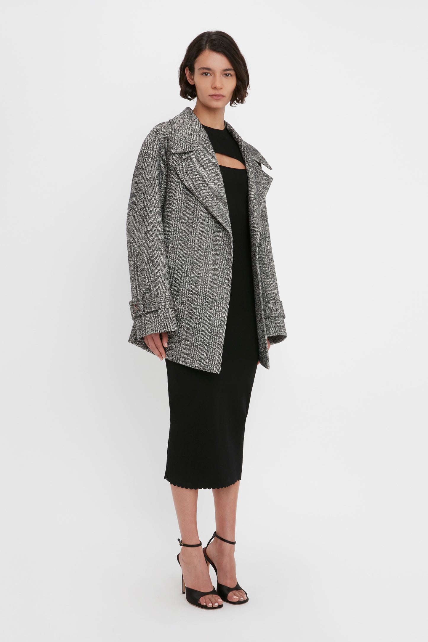 A person stands against a plain background, wearing a gray oversized coat over the Victoria Beckham VB Body Cut Out Midi Dress in Black and black high-heeled sandals.