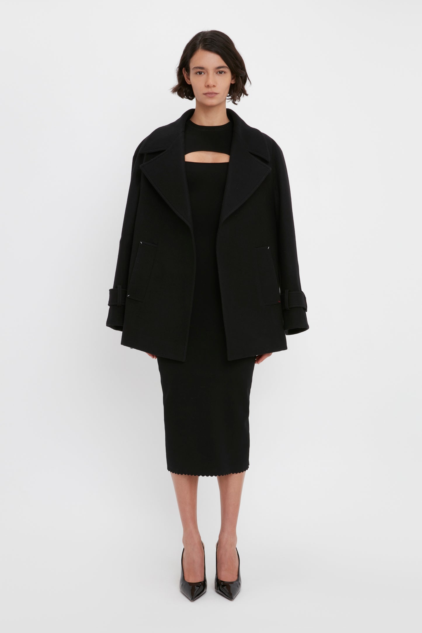 A woman stands against a plain white background wearing a black coat, the VB Body Cut Out Midi Dress in Black by Victoria Beckham, and black high heels.