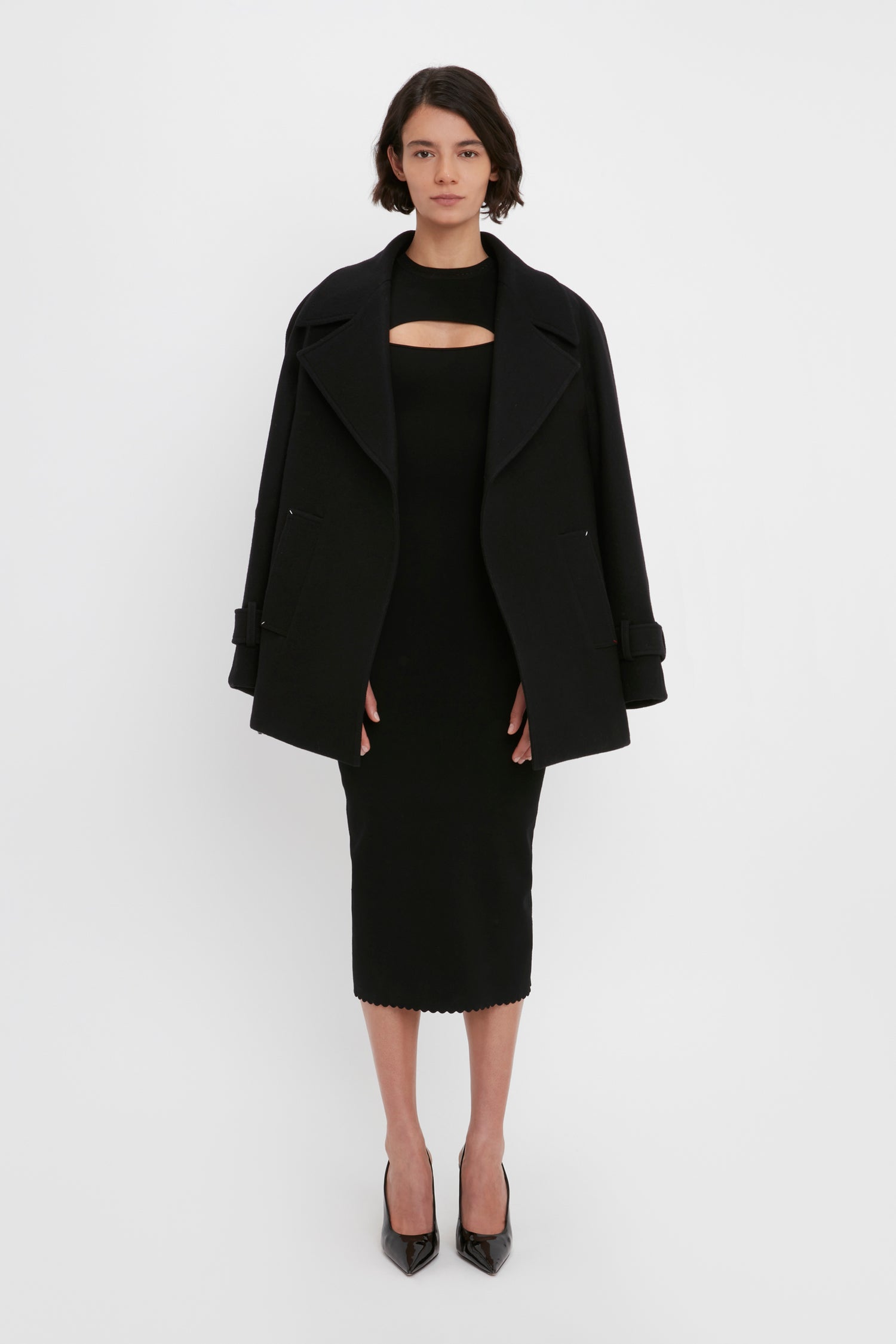 A person stands against a white background, wearing the VB Body Cut Out Midi Dress in Black from Victoria Beckham with a black jacket draped over their shoulders. They are also wearing black heels.