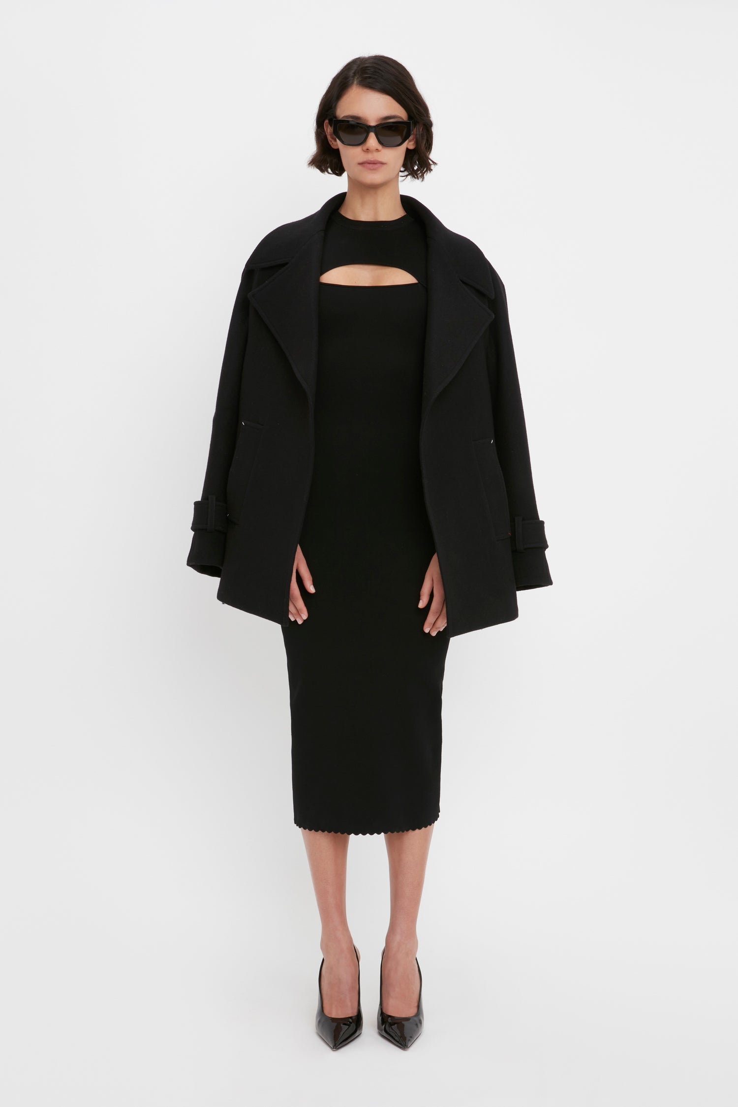 A woman in a VB Body Cut Out Midi Dress by Victoria Beckham, paired with a black oversized coat, black high heels, and sunglasses, standing against a plain white background.
