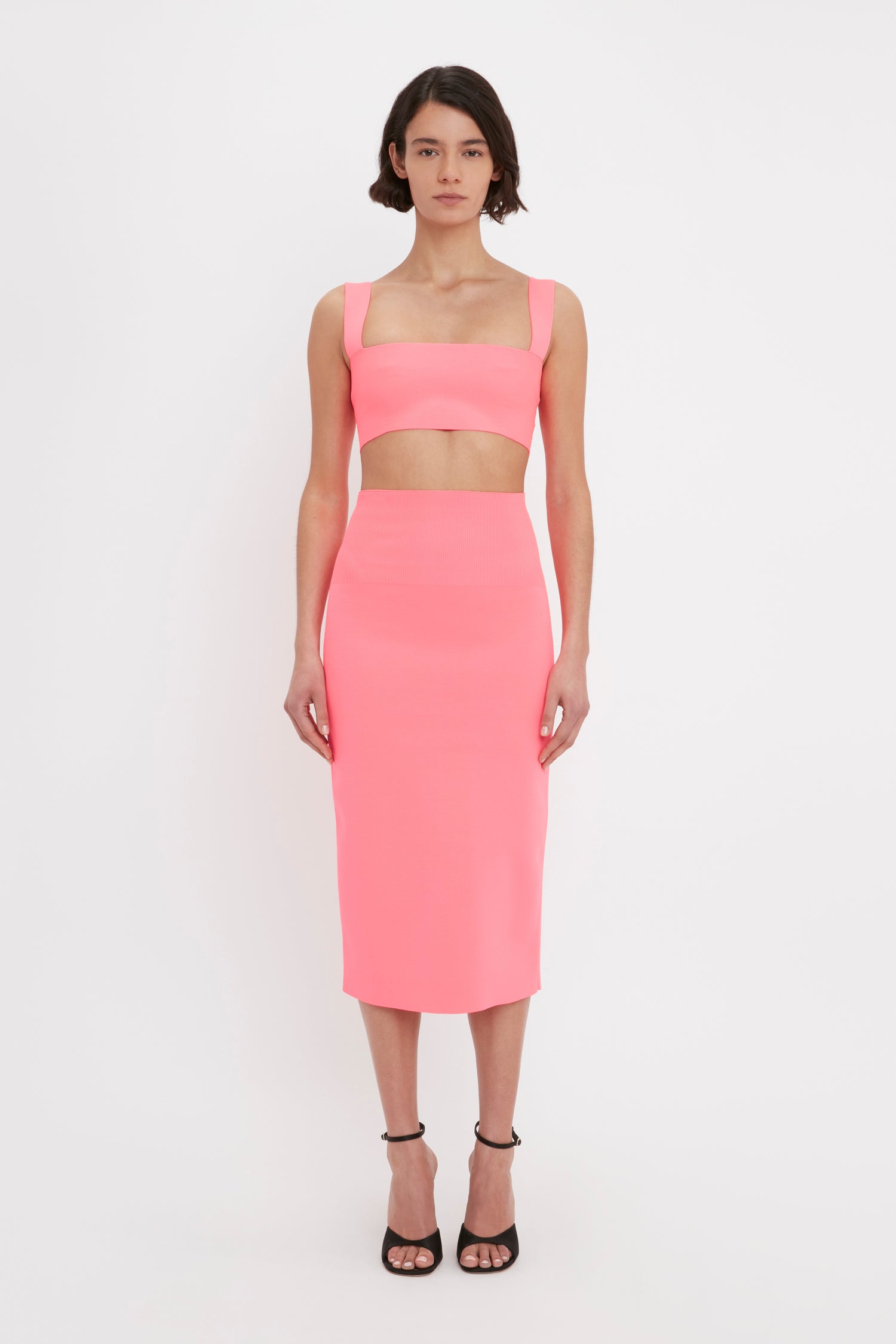 Person in a Victoria Beckham VB Body Strap Bandeau Top in pink and matching high-waisted midi skirt stands against a plain white background. The person is wearing black heeled sandals.