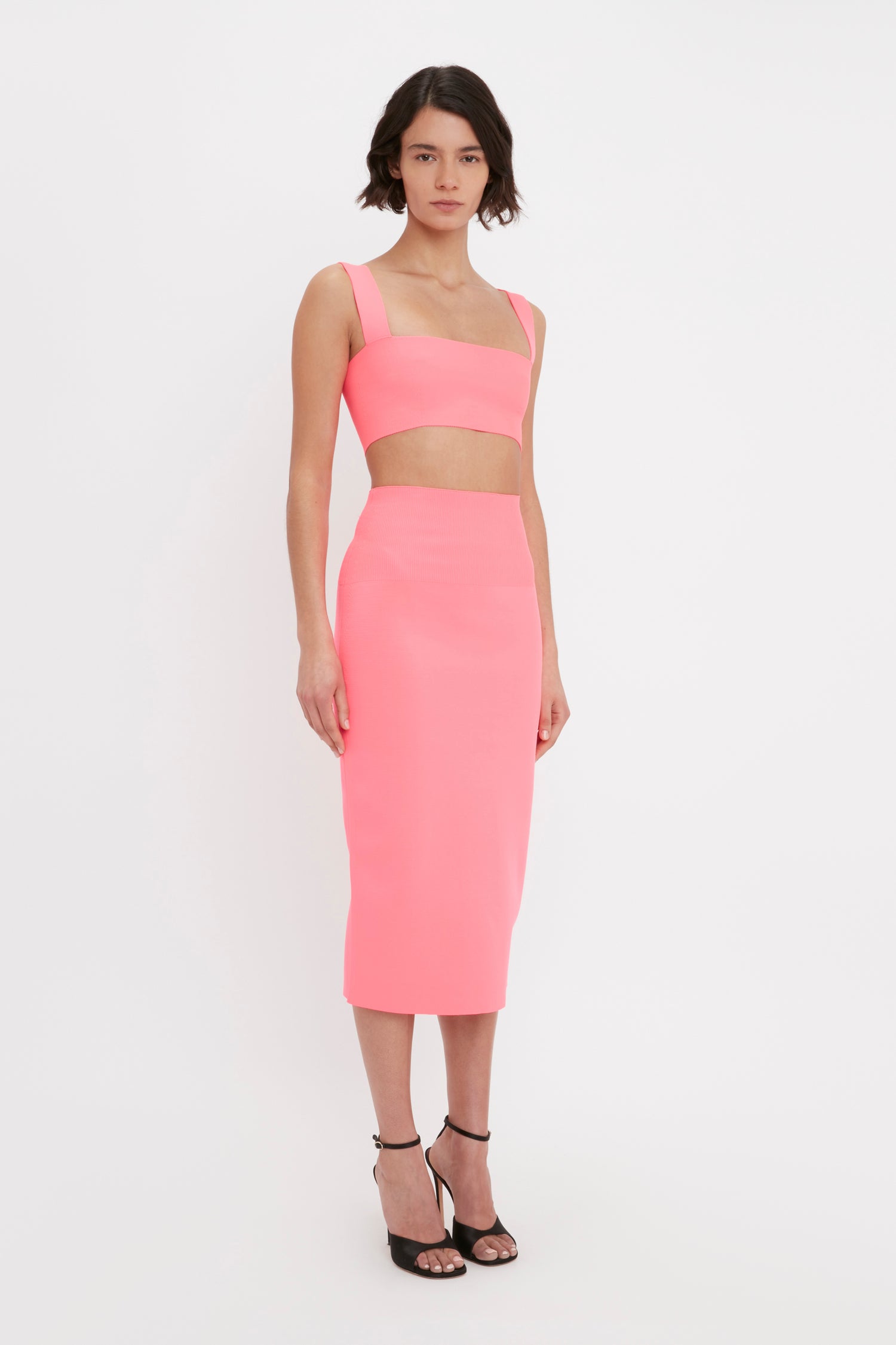 A person stands against a plain white background wearing the Victoria Beckham VB Body Fitted Midi Skirt in Pink, accompanied by a matching crop top and black high-heeled shoes.