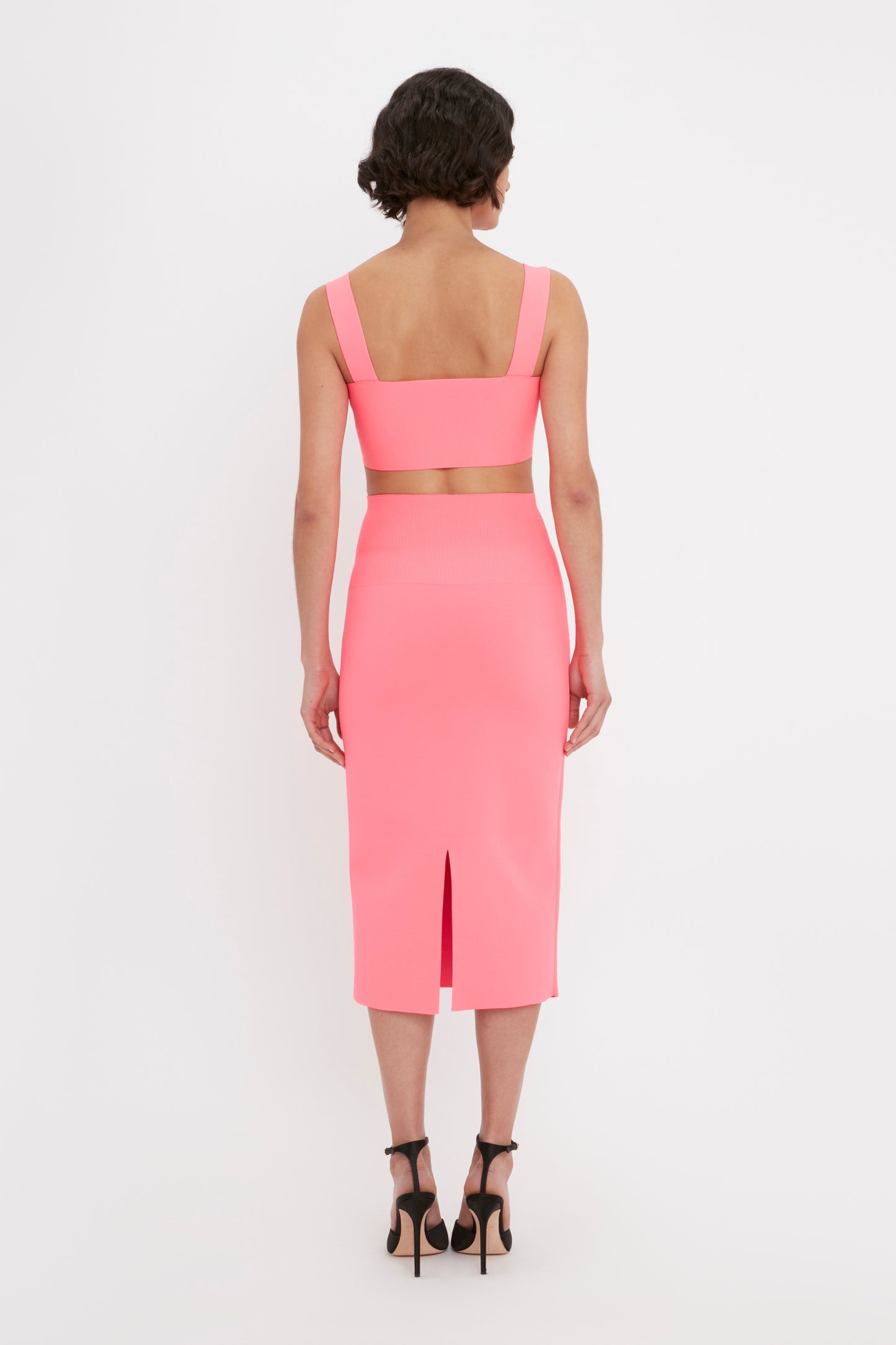 A person with short hair stands facing away, wearing the VB Body Strap Bandeau Top in pink by Victoria Beckham, paired with a matching high-waisted skirt with a back slit, and high heels.