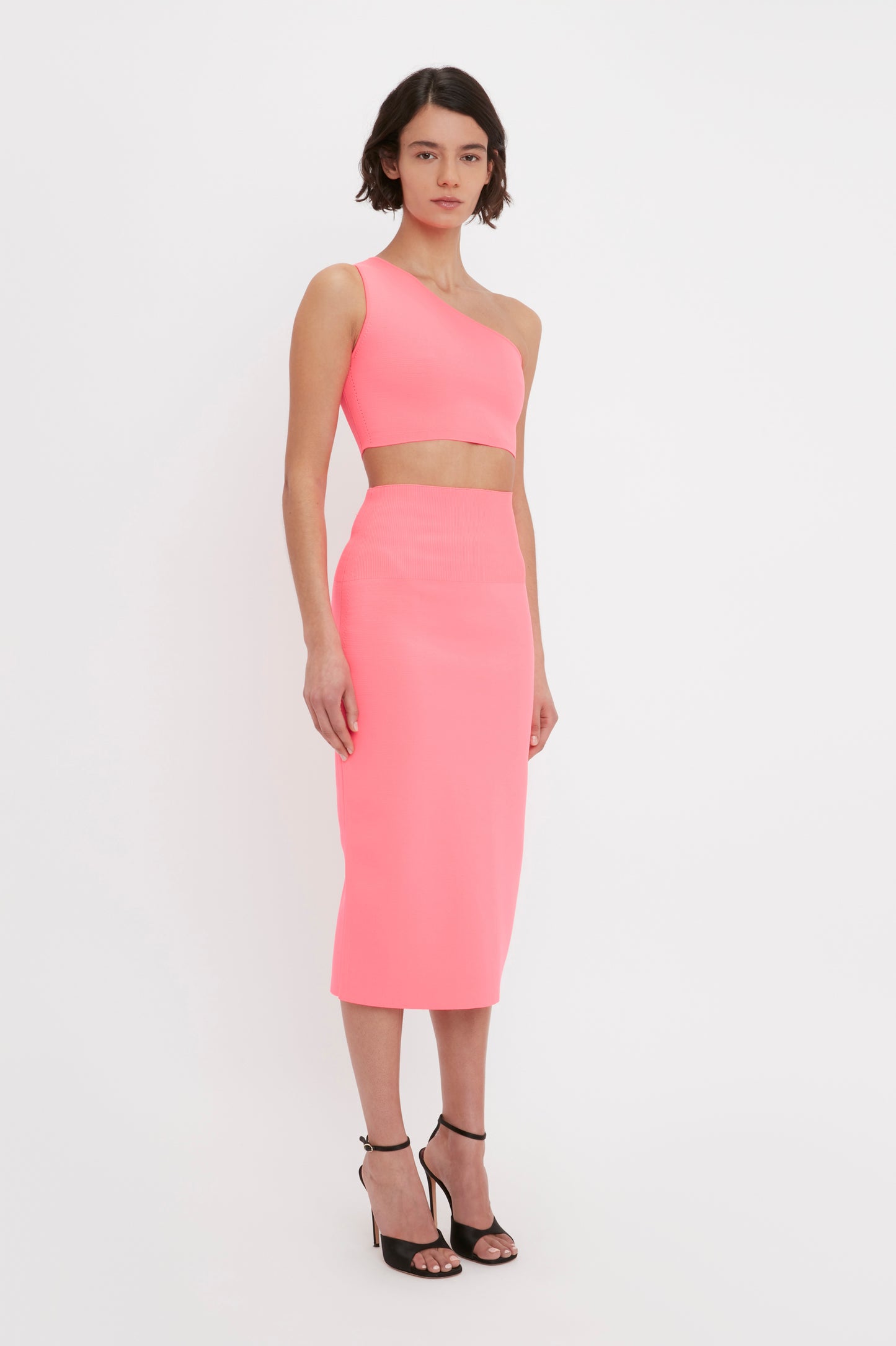 A woman is standing against a white background, wearing the Victoria Beckham VB Body One Shoulder Crop Top in Pink with a matching high-waisted, knee-length skirt, paired with black high-heeled sandals.