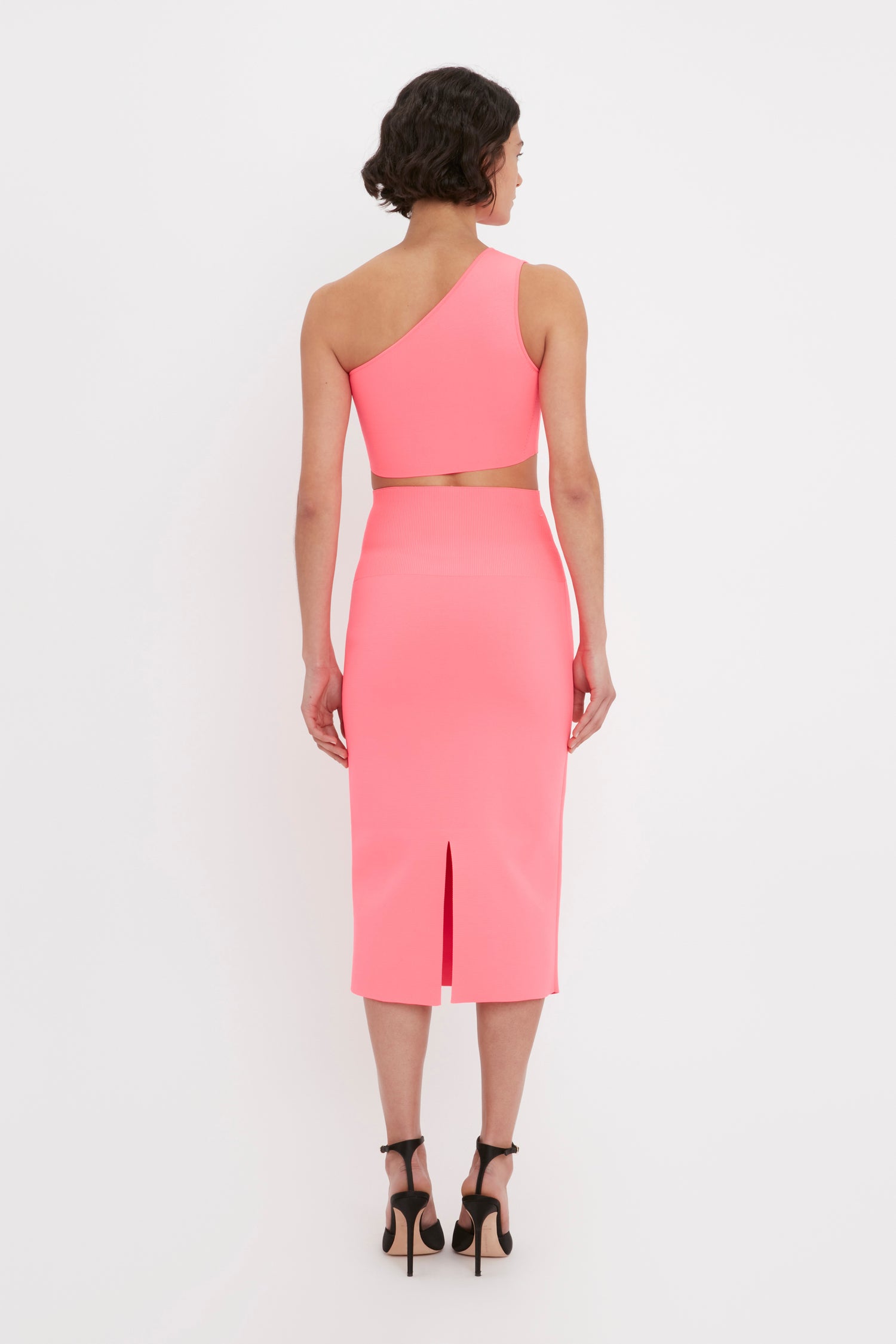 A woman with short dark hair, wearing the Victoria Beckham VB Body One Shoulder Crop Top in pink and black high heels, is standing facing away from the camera.