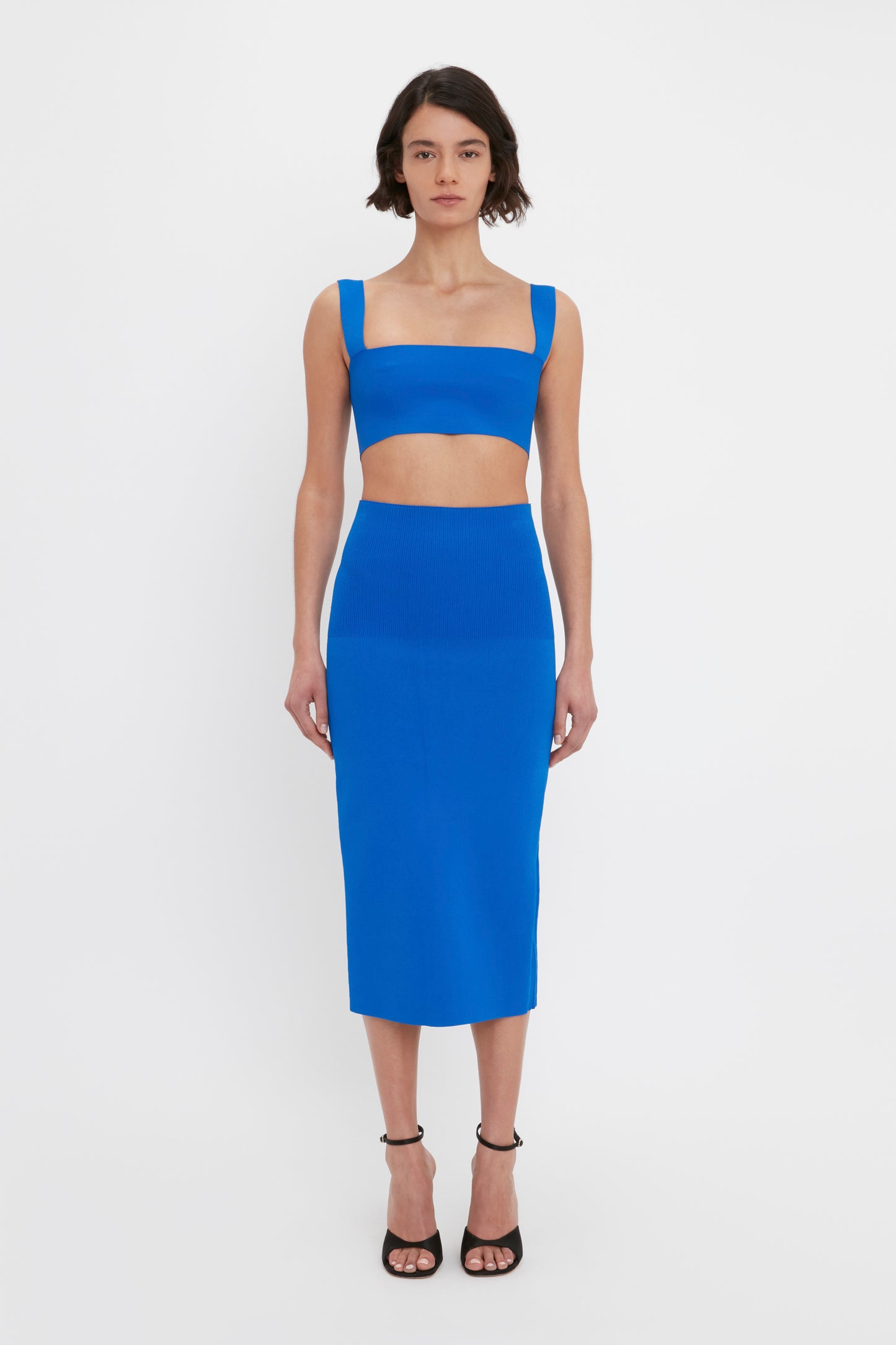 A woman with short brown hair is wearing the Victoria Beckham VB Body Strap Bandeau Top in Cobalt and a matching high-waisted, knee-length skirt, posing against a plain white background. She is also wearing black ankle-strap heels.