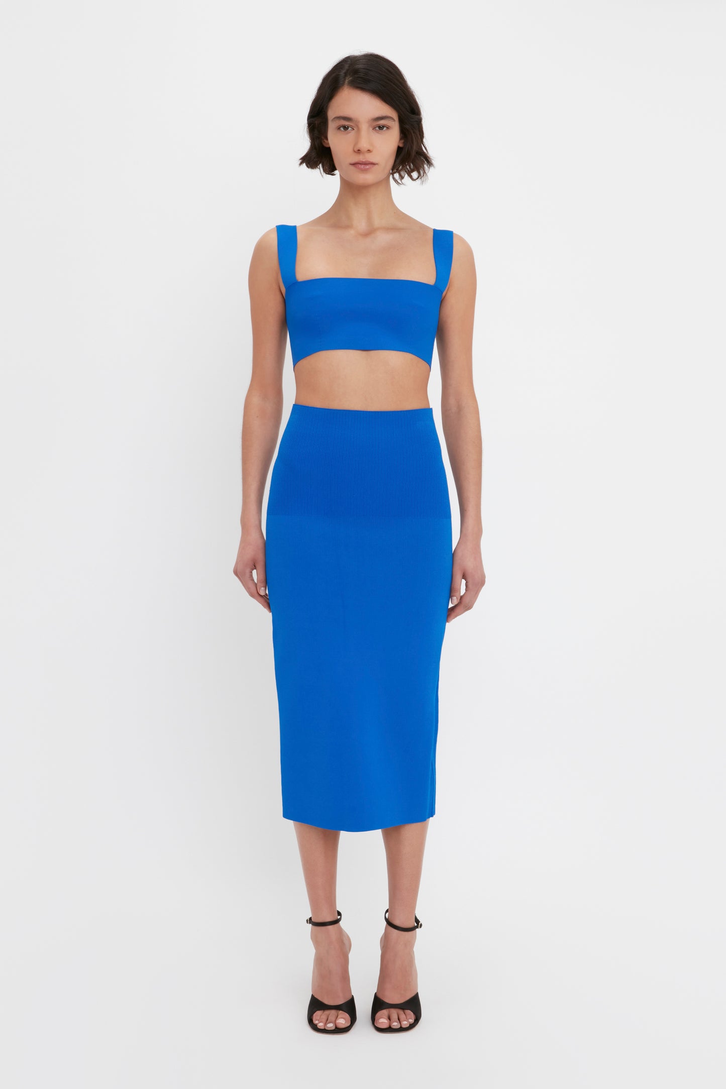 A person stands against a white background, wearing a cobalt blue crop top and the VB Body Fitted Midi Skirt in Cobalt by Victoria Beckham, paired with black high-heeled sandals.