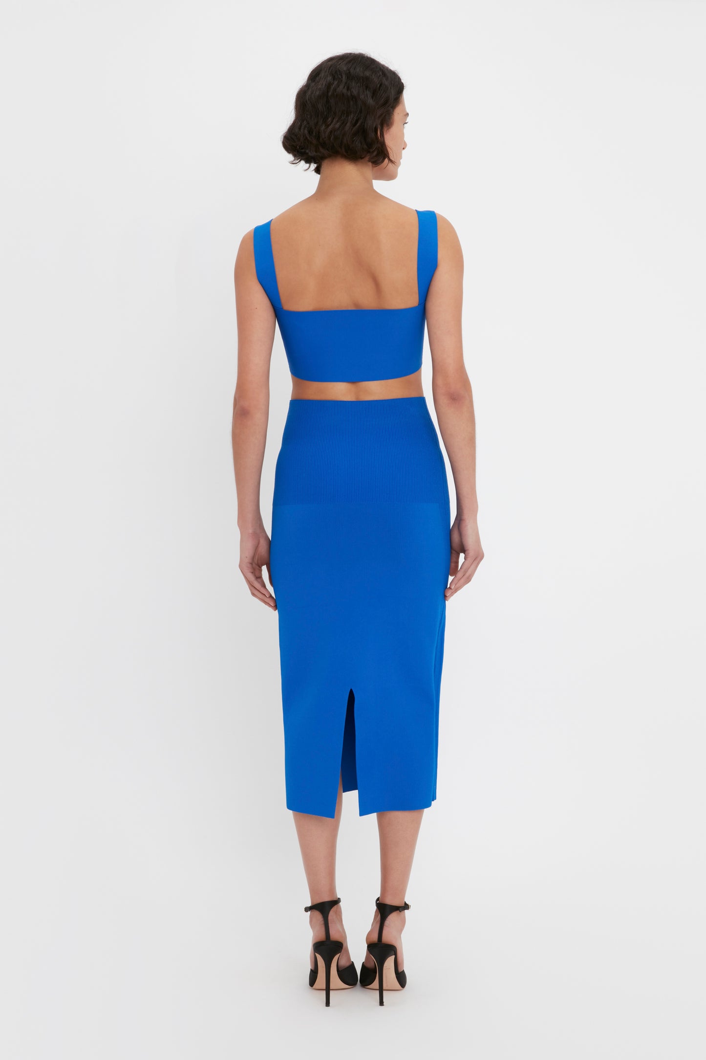 A person with short dark hair is wearing the Victoria Beckham VB Body Fitted Midi Skirt in Colbalt, designed with an open back and a slit. They are standing facing away from the camera against a white background.