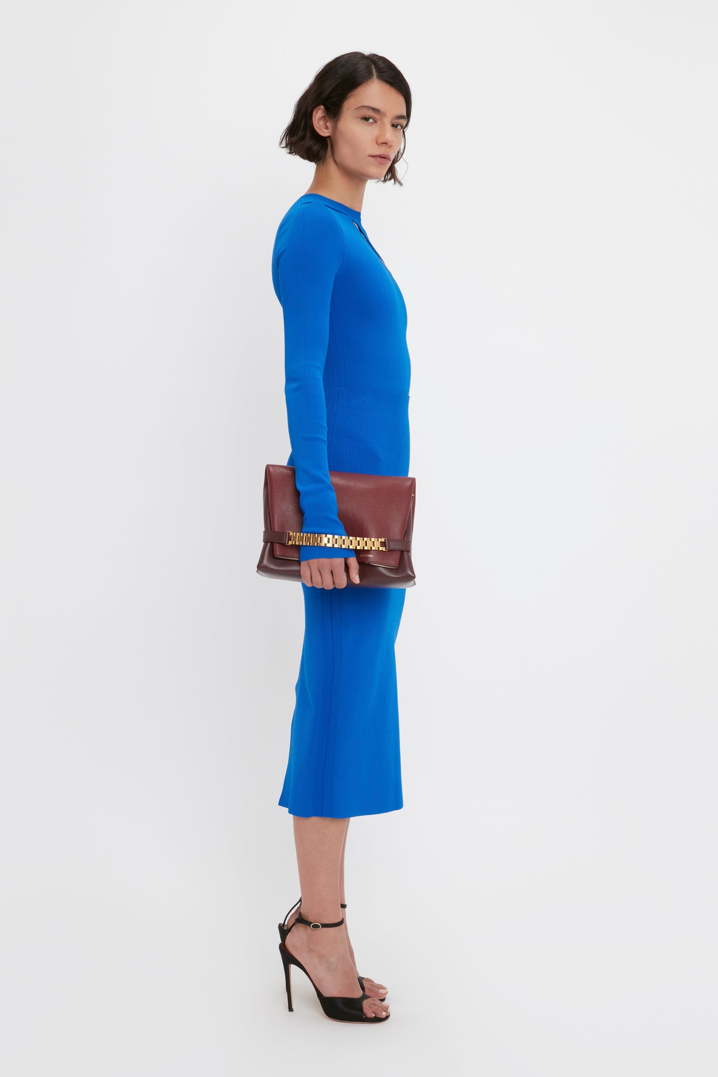A person stands in profile wearing the VB Body Front Slash Crew Neck Top in Cobalt by Victoria Beckham and black high heels, holding a brown clutch with a decorative strap. The background is plain white.