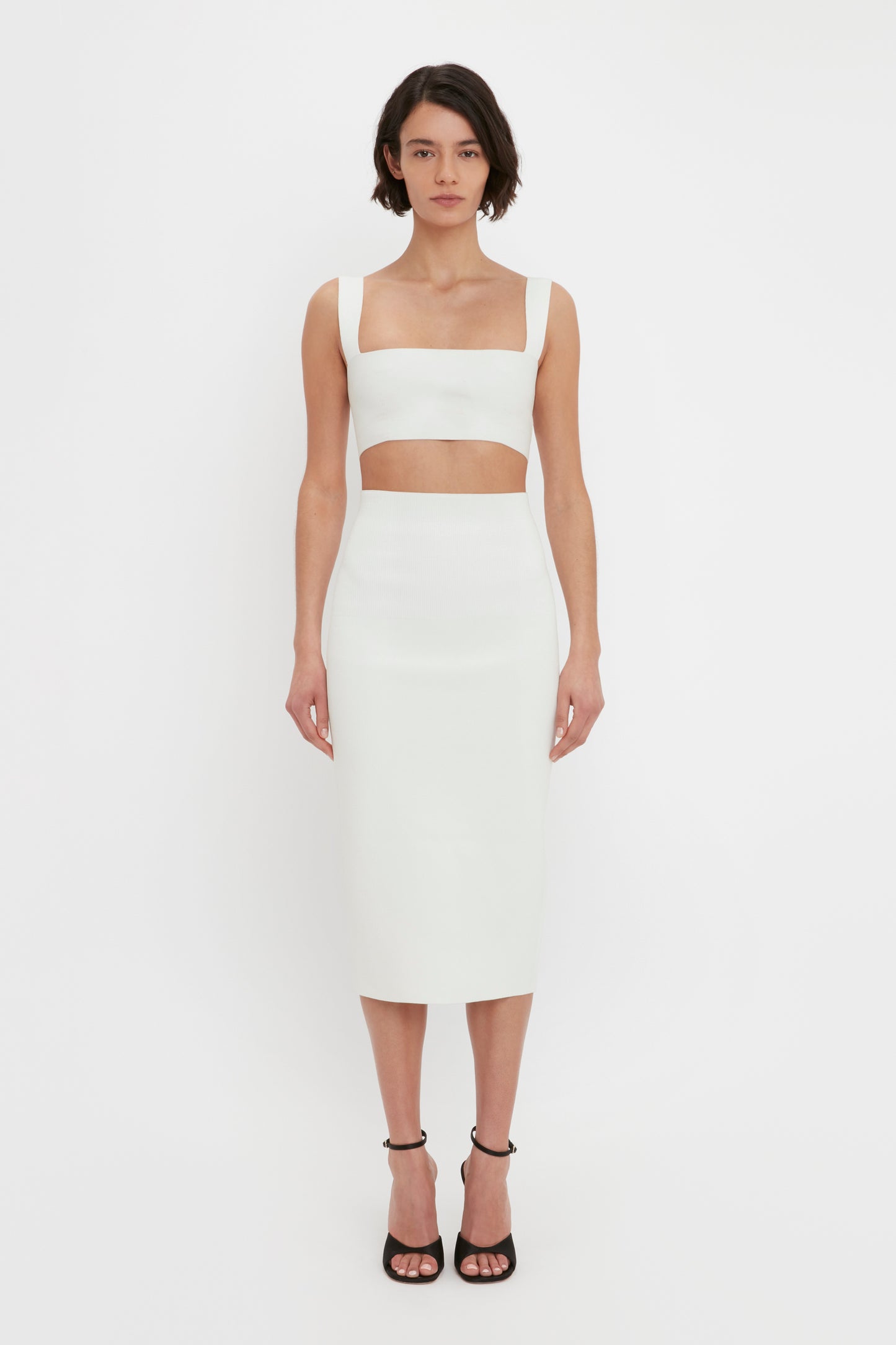 A person stands against a plain white background, wearing the VB Body Strap Bandeau Top in White by Victoria Beckham and a matching high-waisted skirt extending below the knees, paired with black high-heeled shoes.