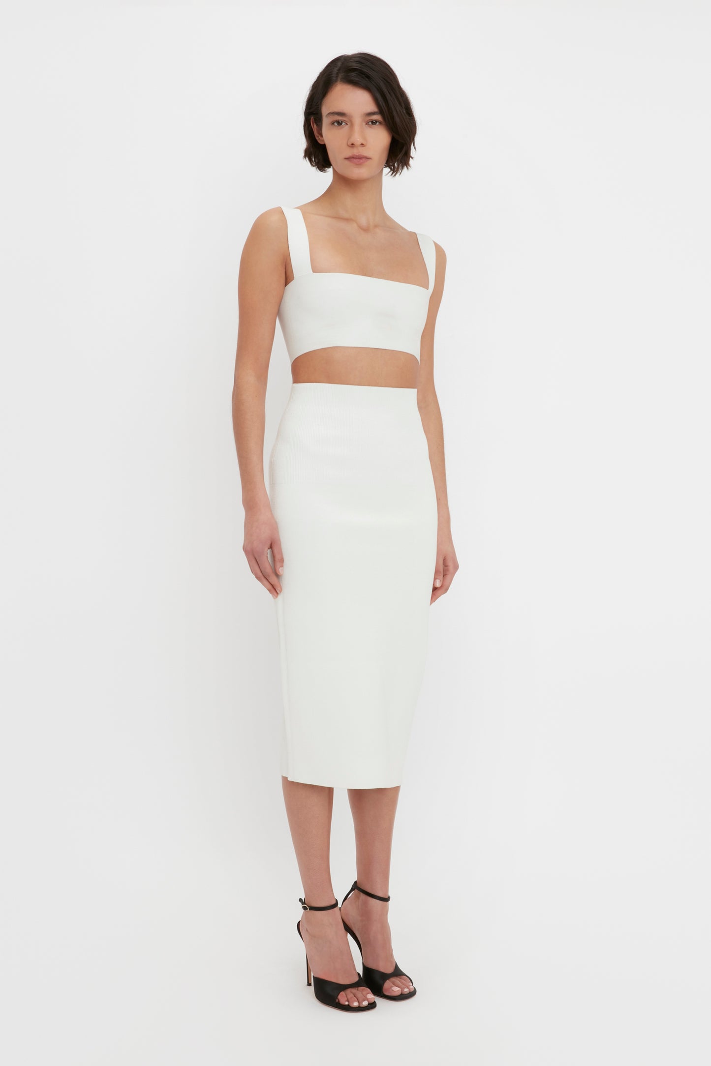A woman in a white VB Body Strap Bandeau Top from Victoria Beckham and a matching midi skirt stands against a plain white background, wearing black high-heeled sandals.