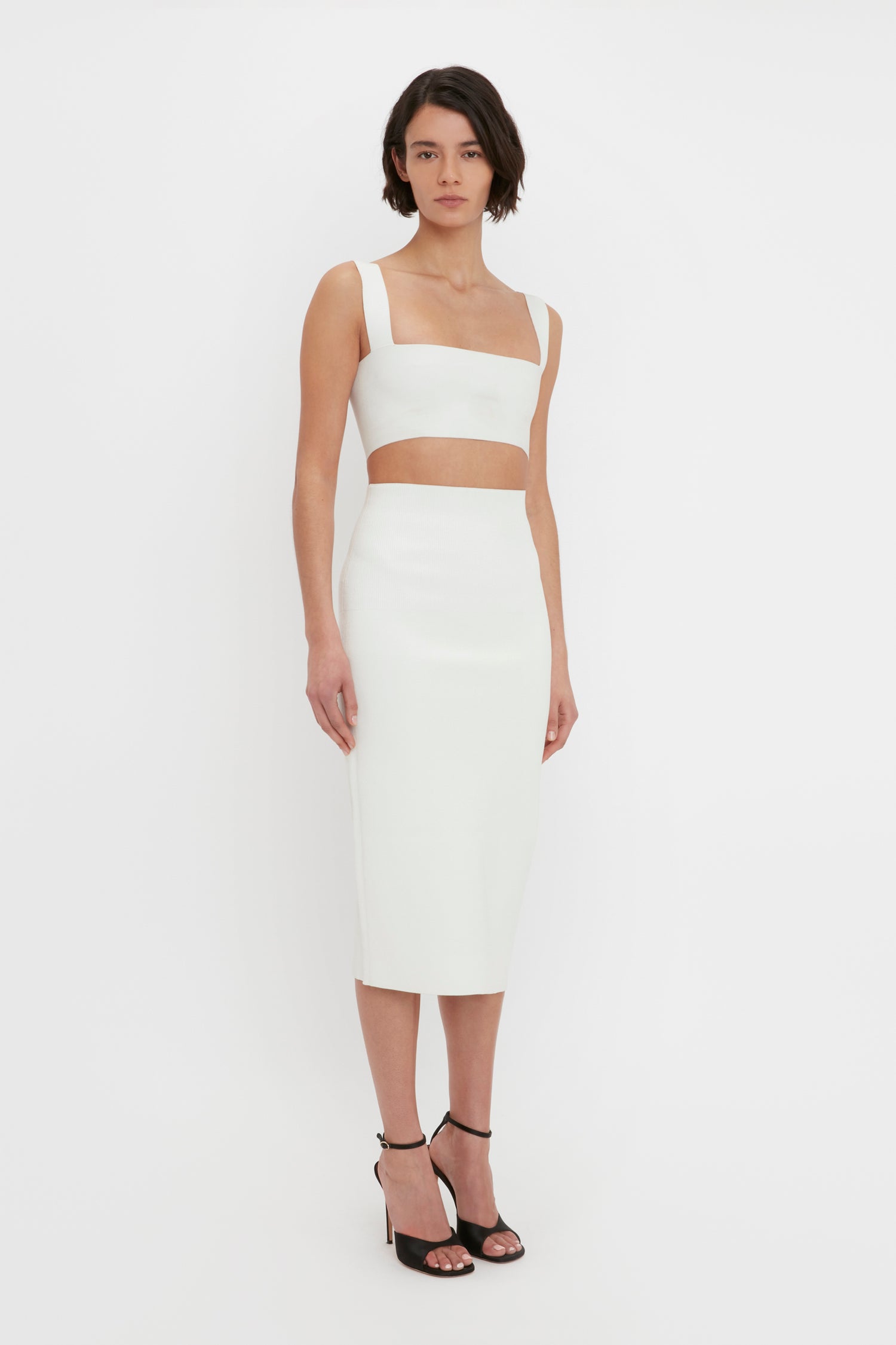 A woman in a white VB Body Strap Bandeau Top from Victoria Beckham and a matching midi skirt stands against a plain white background, wearing black high-heeled sandals.