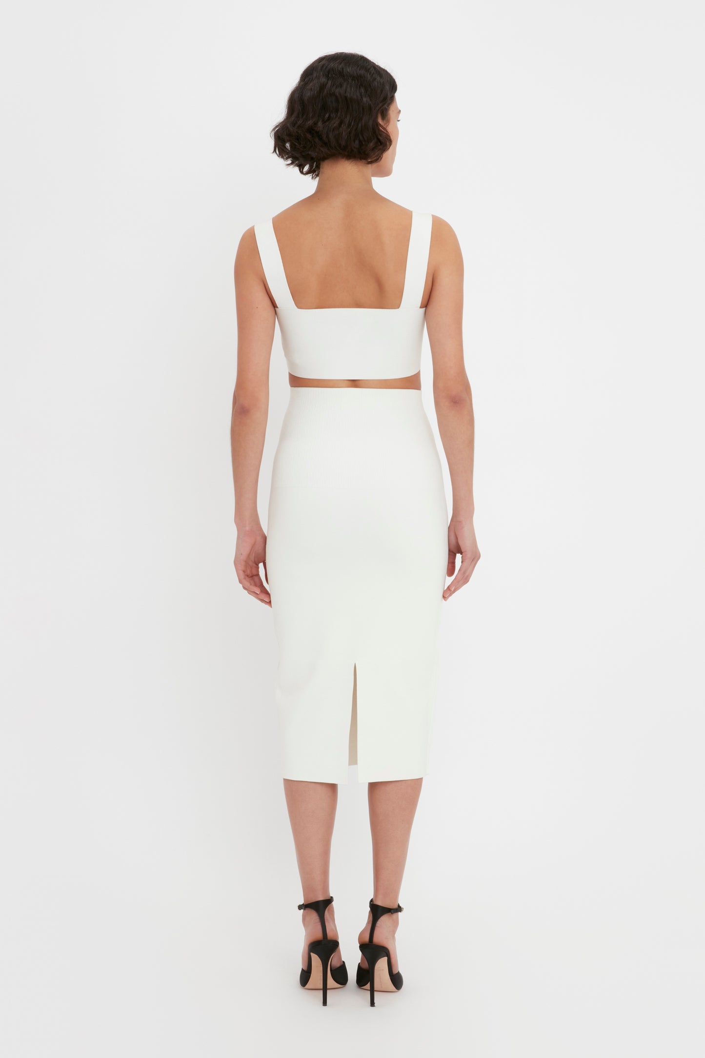 A woman with short curly hair is wearing the VB Body Strap Bandeau Top in White by Victoria Beckham, paired with a coordinating bottom and black heels. She is standing and facing away from the camera, highlighting the back of her outfit.