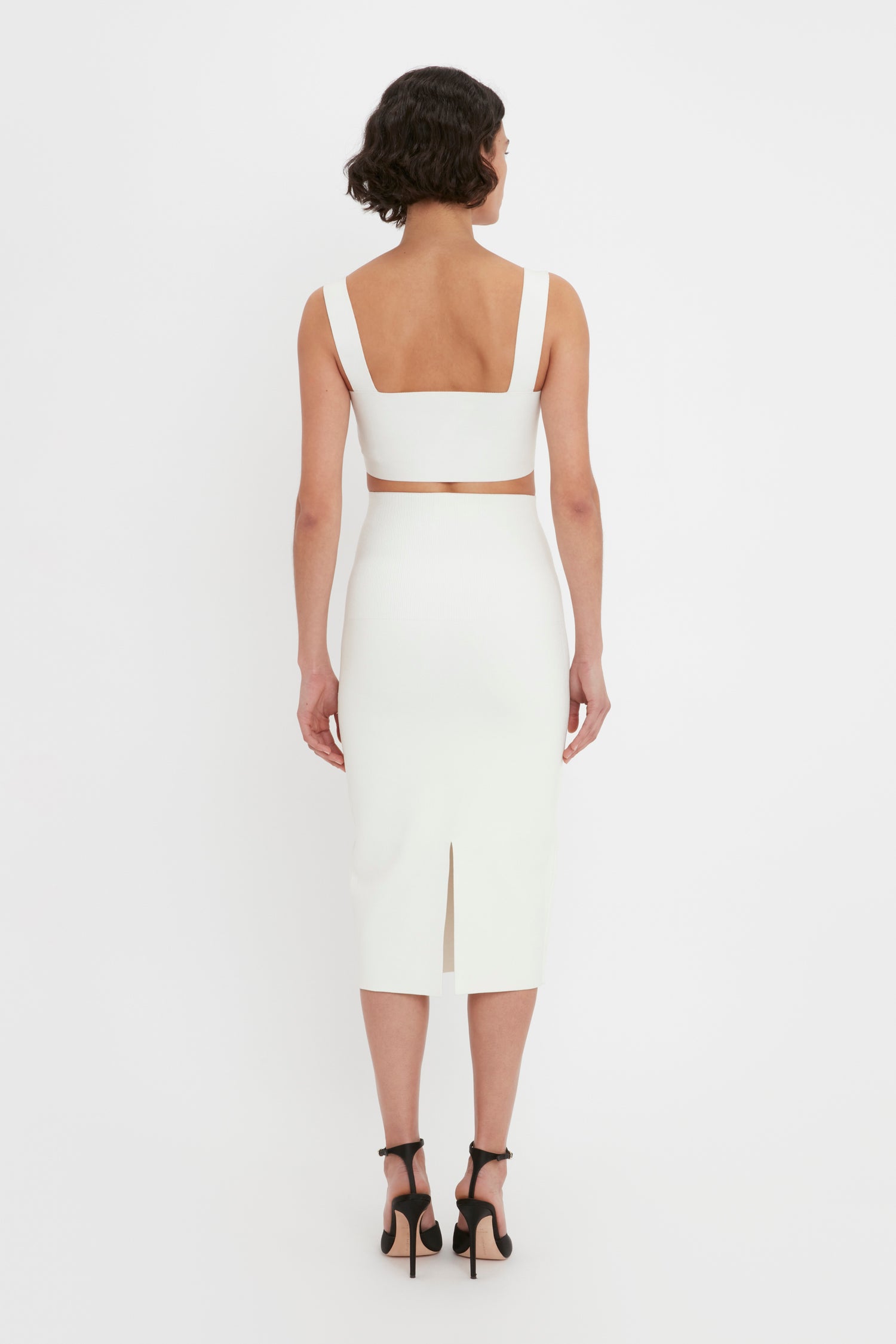 A woman with short curly hair is wearing the VB Body Strap Bandeau Top in White by Victoria Beckham, paired with a coordinating bottom and black heels. She is standing and facing away from the camera, highlighting the back of her outfit.