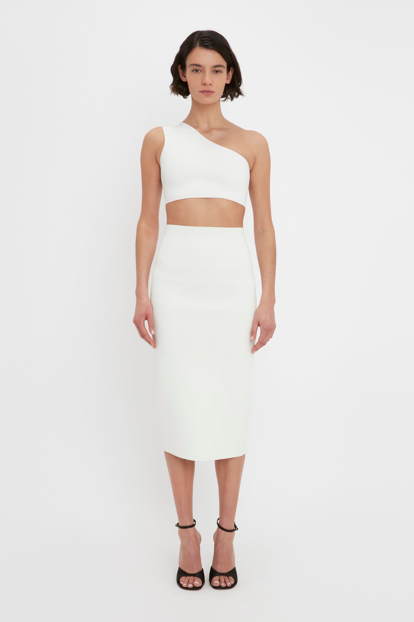 A woman stands against a white background wearing the VB Body One Shoulder Crop Top in White by Victoria Beckham, paired with a matching high-waisted pencil skirt and black peep-toe heels.