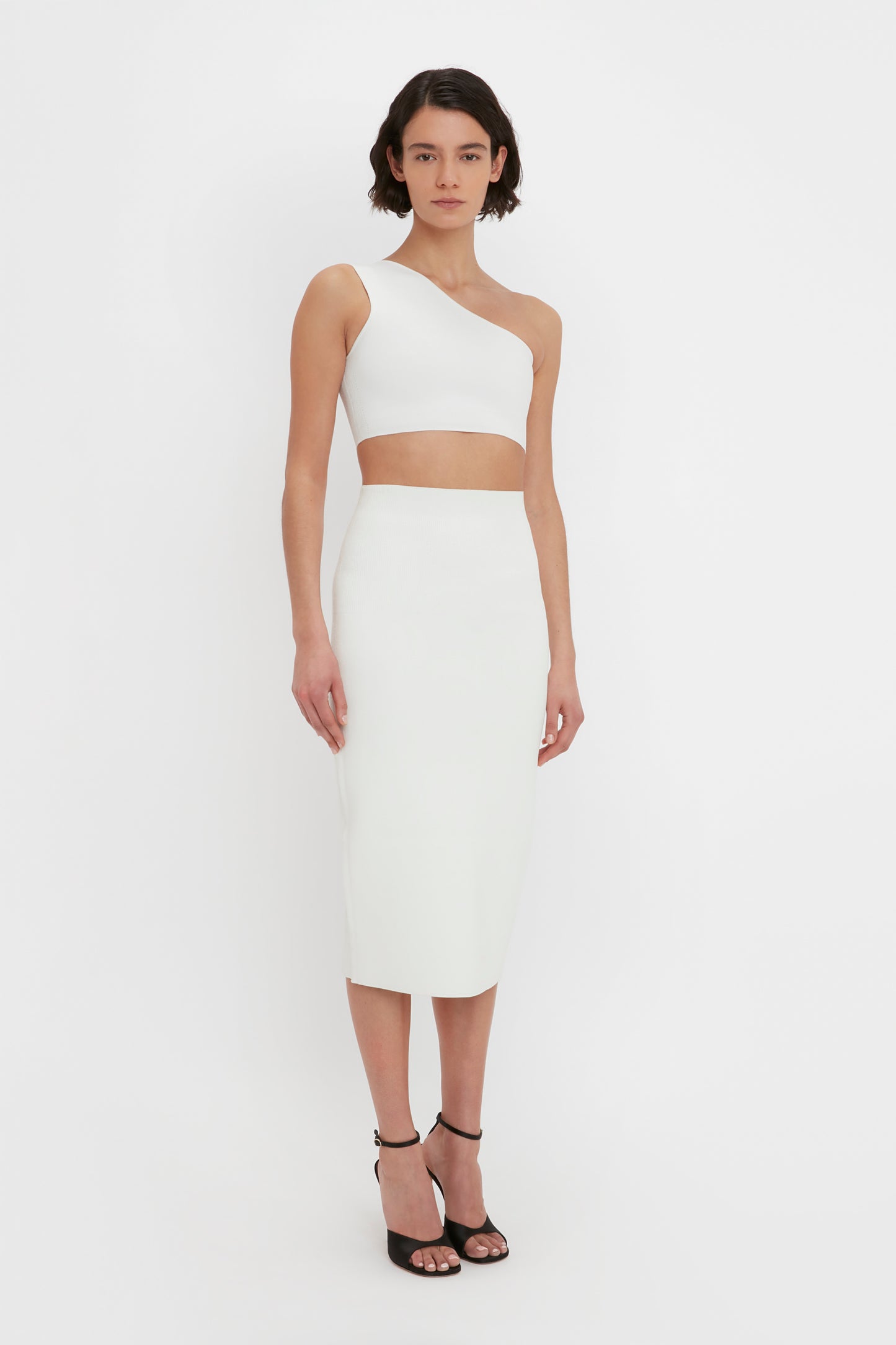 A person with short hair wears a white one-shoulder crop top and the VB Body Fitted Midi Skirt in White by Victoria Beckham, paired with black high-heeled sandals, standing against a plain white background.