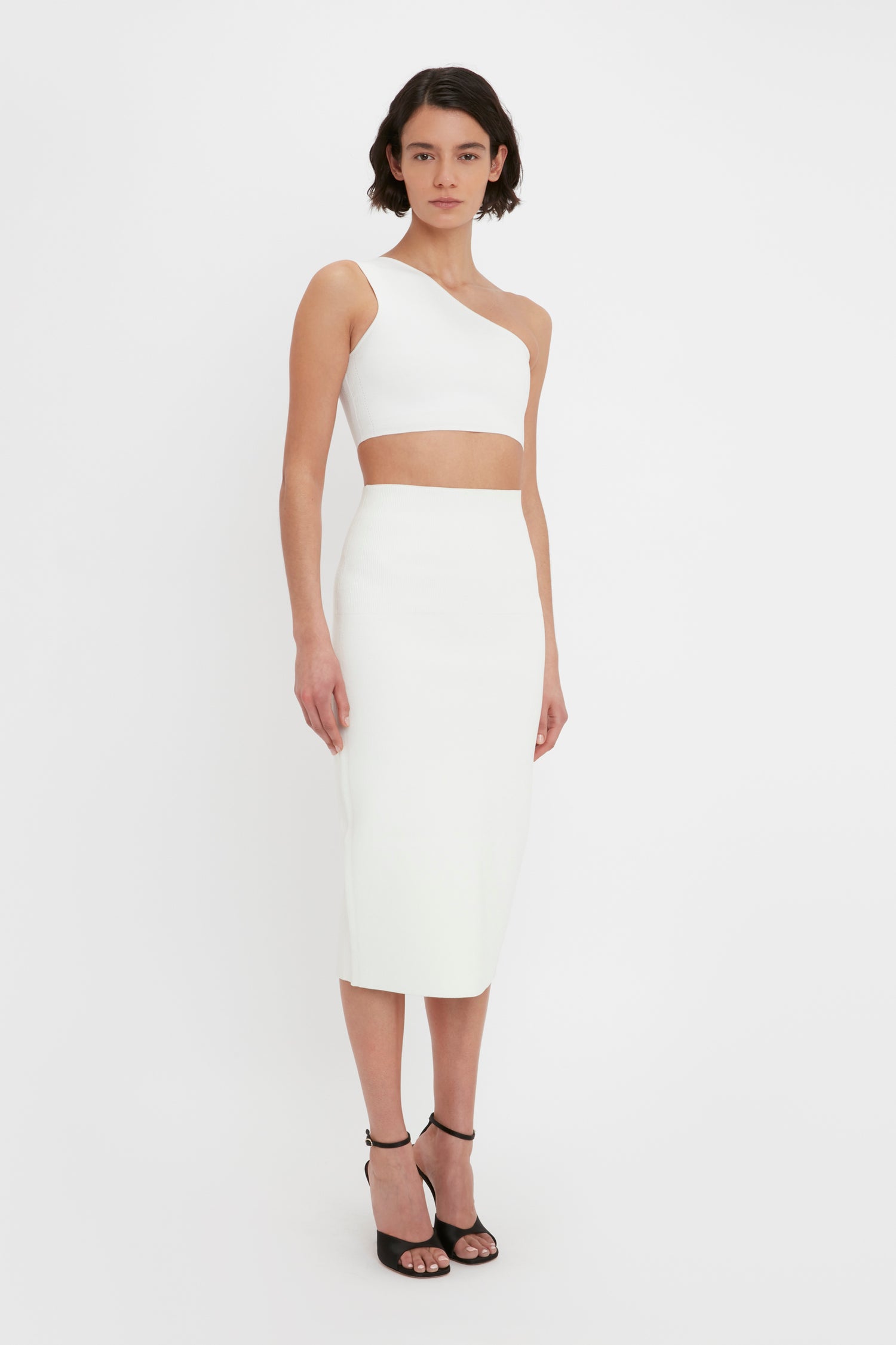 Person wearing a VB Body One Shoulder Crop Top in White by Victoria Beckham and a matching white high-waisted, knee-length skirt, paired with black high-heeled sandals.