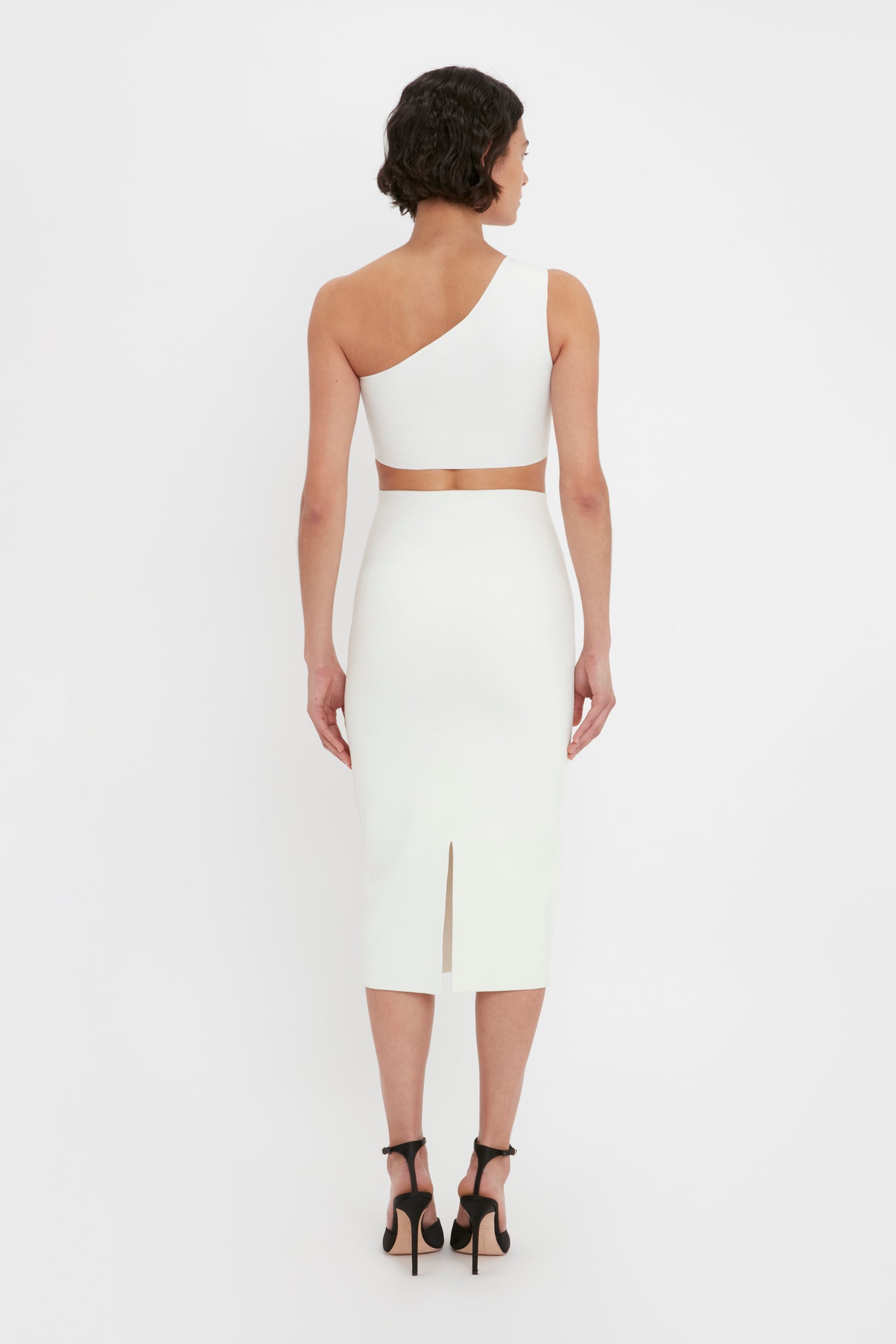 A woman stands with her back facing the camera, wearing the VB Body Fitted Midi Skirt in White by Victoria Beckham. She has short, dark hair and is wearing black high-heeled shoes.