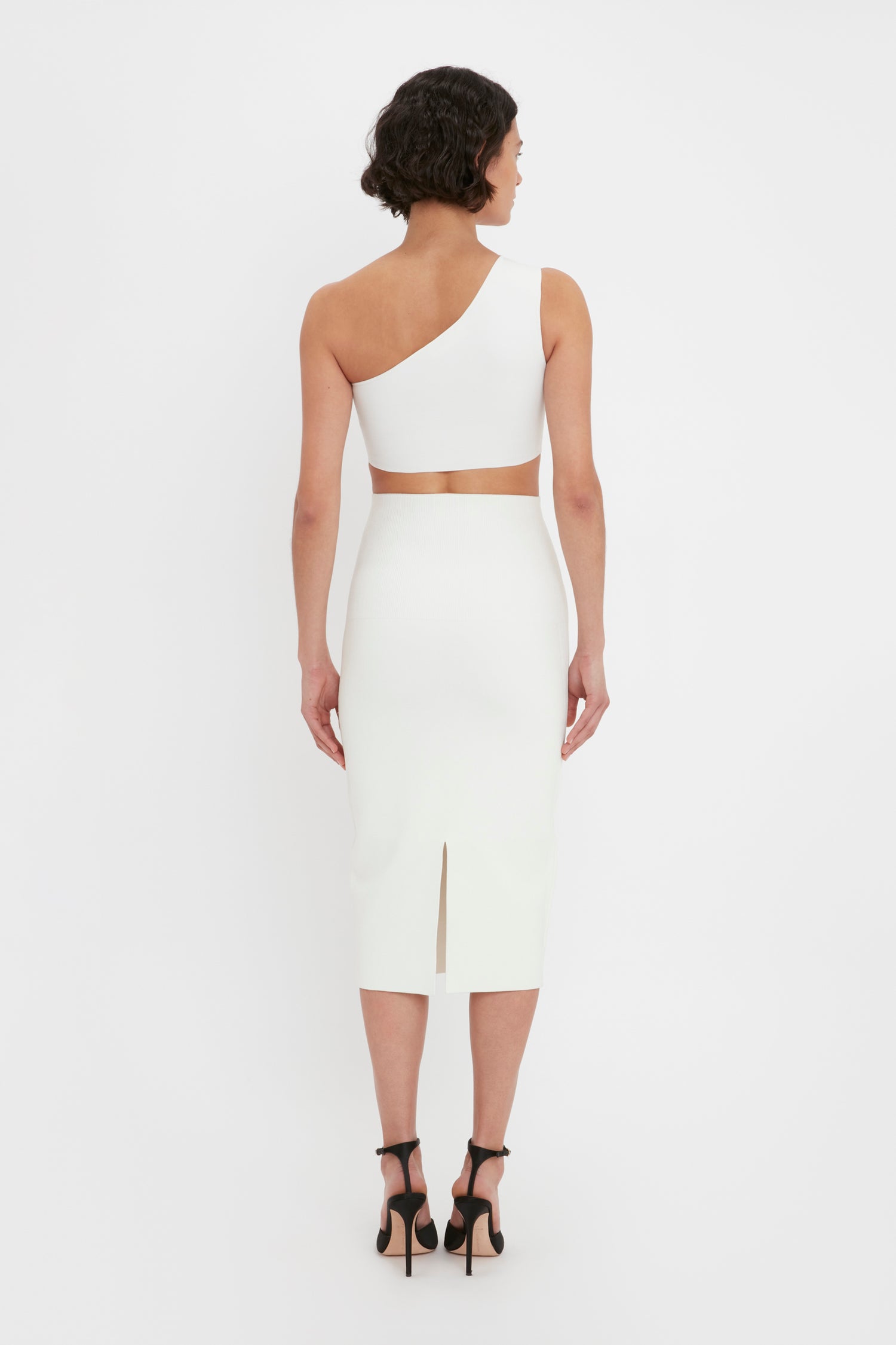 A woman with short dark hair is wearing a Victoria Beckham VB Body One Shoulder Crop Top in white, paired with black high-heeled shoes. She is standing with her back to the camera against a plain white background.