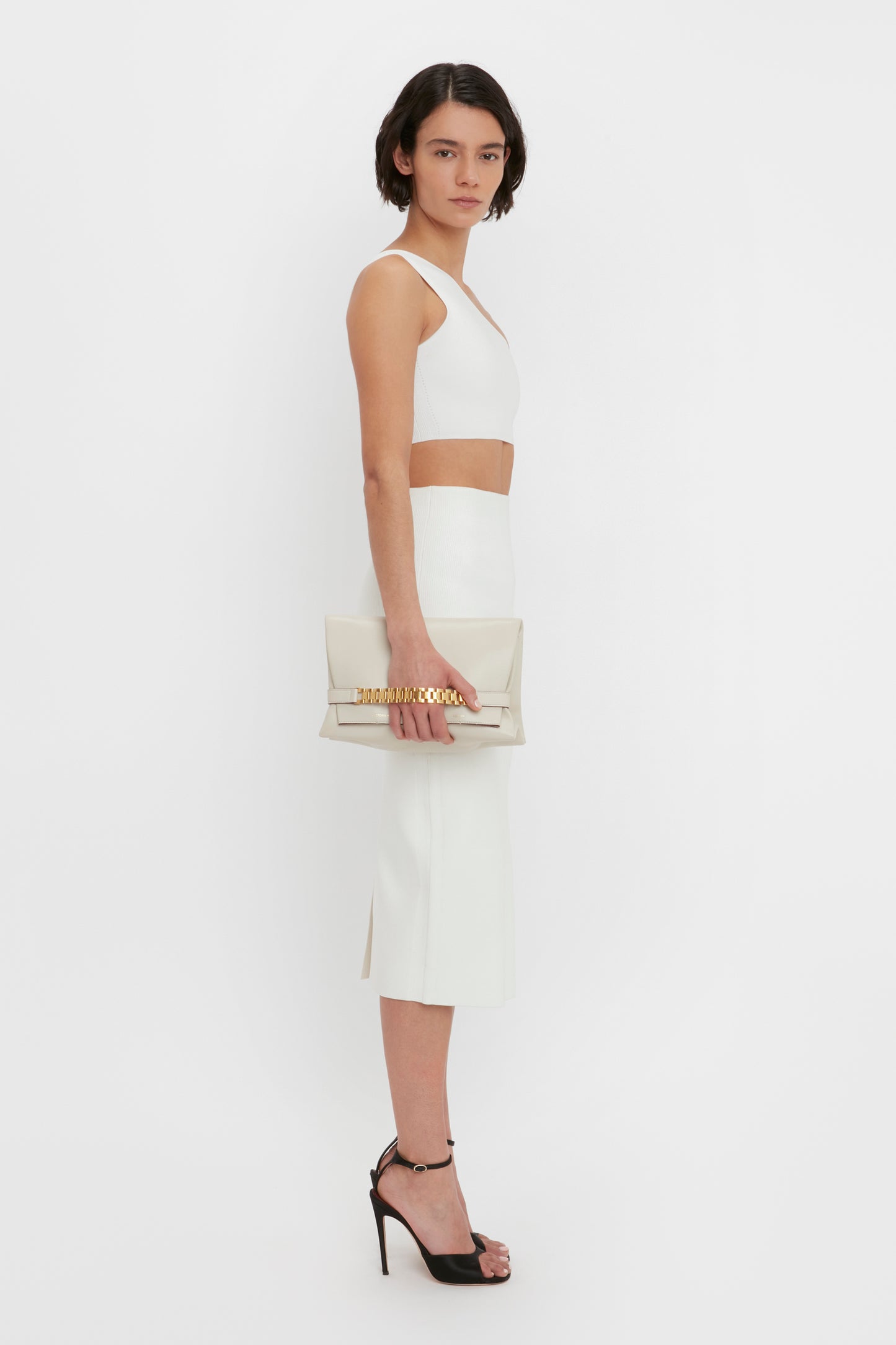 A person stands facing forward and looking to their right against a plain white background, wearing the VB Body Fitted Midi Skirt in White by Victoria Beckham paired with black high heels and holding a beige clutch.