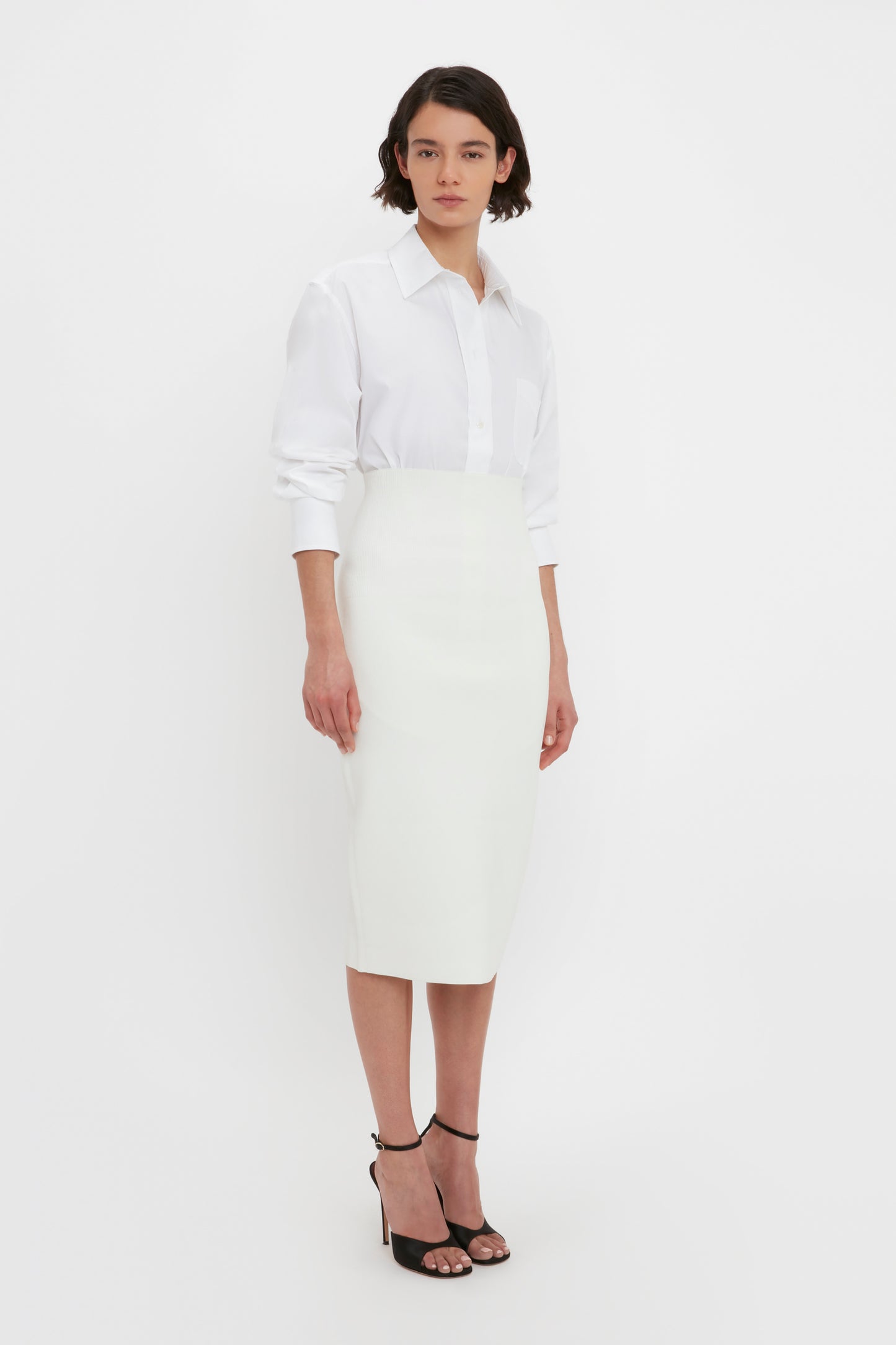 A person with shoulder-length hair is wearing a white shirt and the VB Body Fitted Midi Skirt in White by Victoria Beckham, standing against a plain background.