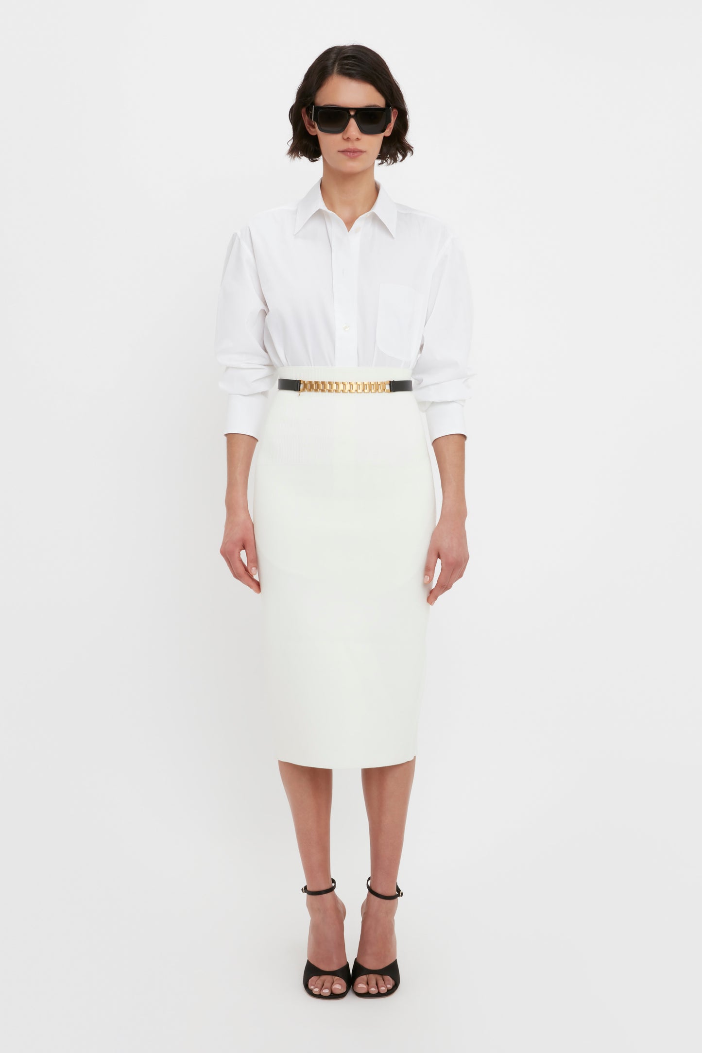 A person wearing a white blouse, the VB Body Fitted Midi Skirt in White by Victoria Beckham, a black belt with a gold chain detail, black high-heel sandals, and sunglasses stands against a plain white background.