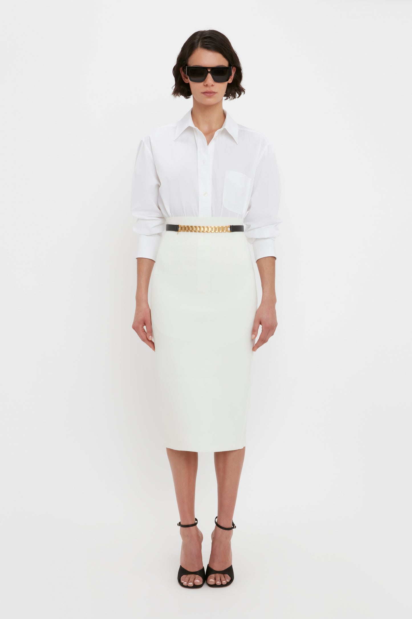 A woman stands against a white background wearing sunglasses, a white collared shirt, the VB Body Fitted Midi Skirt in White by Victoria Beckham, and black high-heeled sandals.
