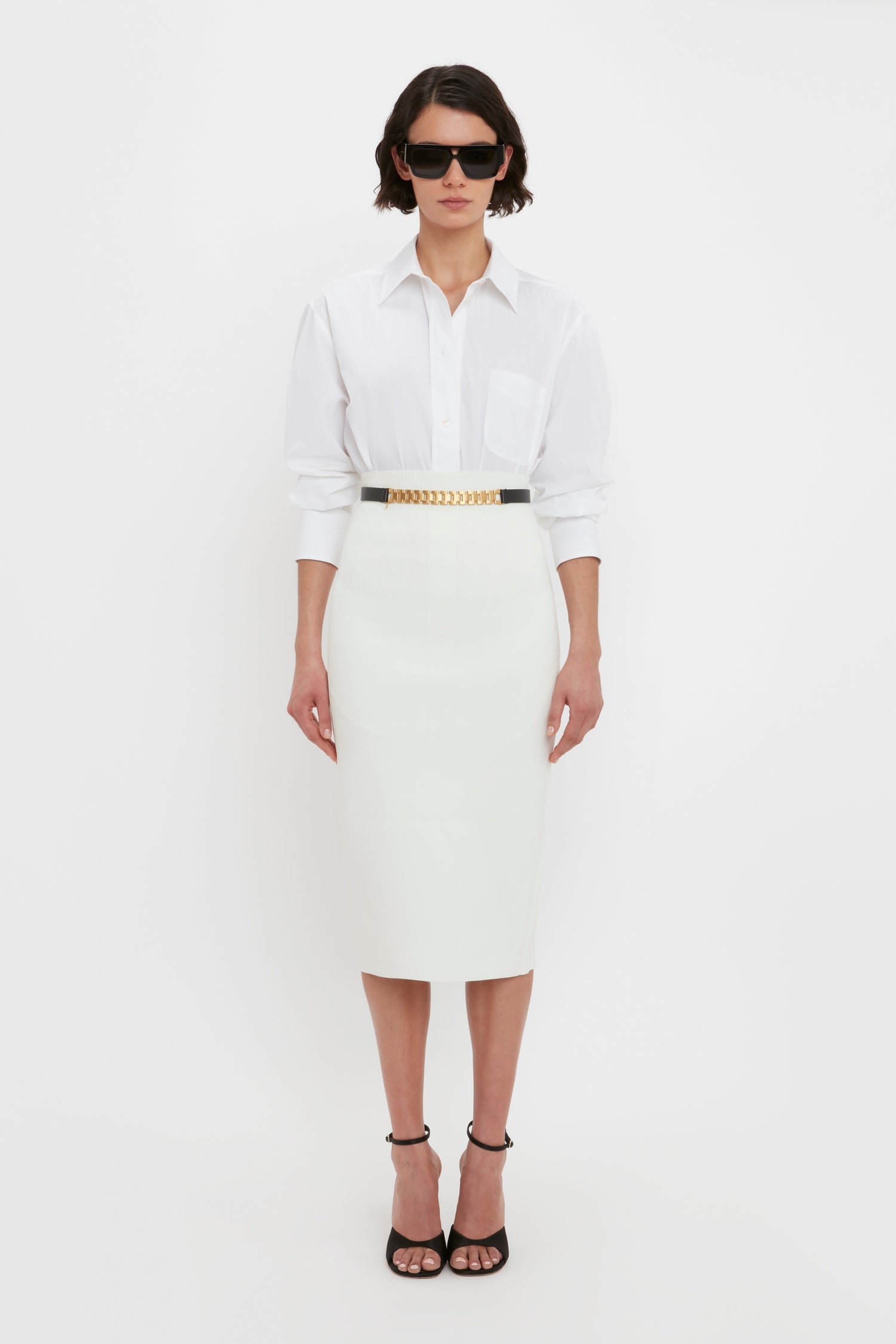 A woman stands against a white background wearing sunglasses, a white collared shirt, the VB Body Fitted Midi Skirt in White by Victoria Beckham, and black high-heeled sandals.