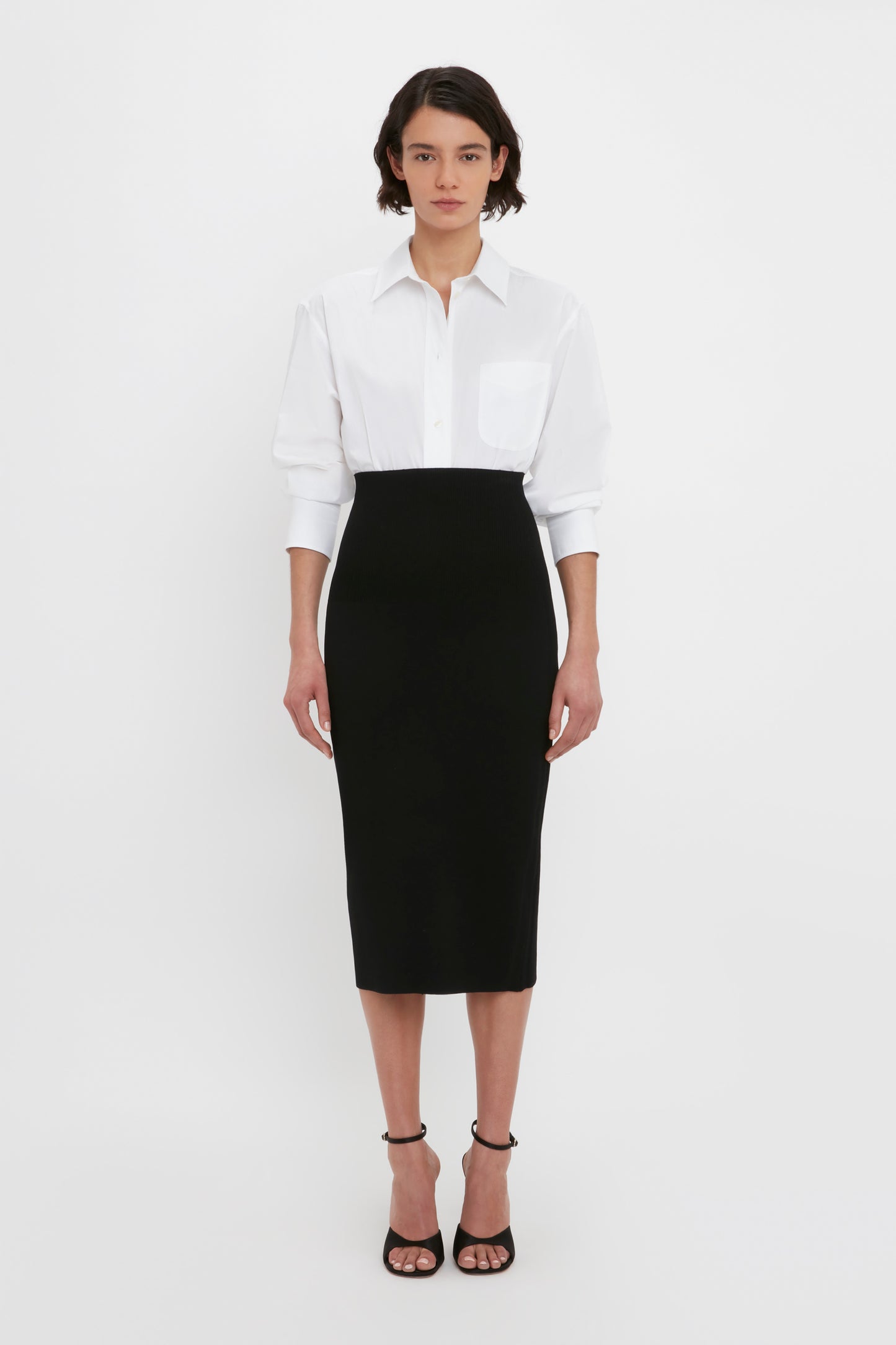 A person stands against a white background, wearing a white collared shirt tucked into the VB Body Fitted Midi Skirt in Black by Victoria Beckham, paired with black open-toe heels.