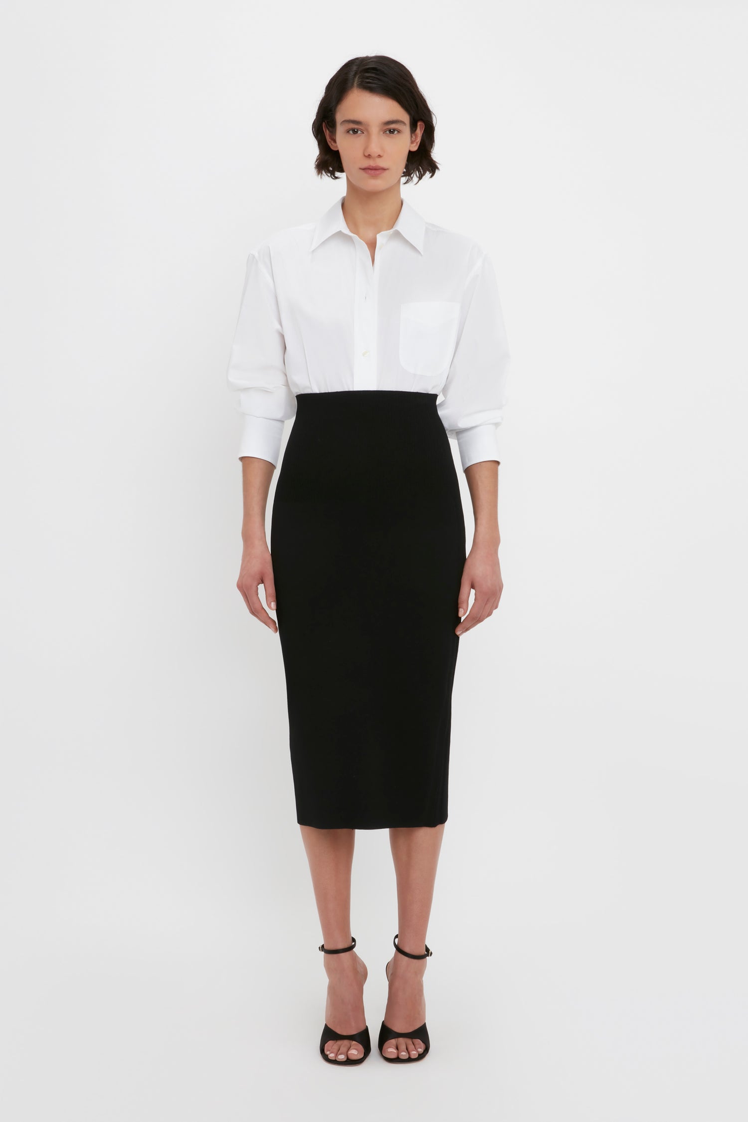 A person stands against a white background, wearing a white collared shirt tucked into the VB Body Fitted Midi Skirt in Black by Victoria Beckham, paired with black open-toe heels.