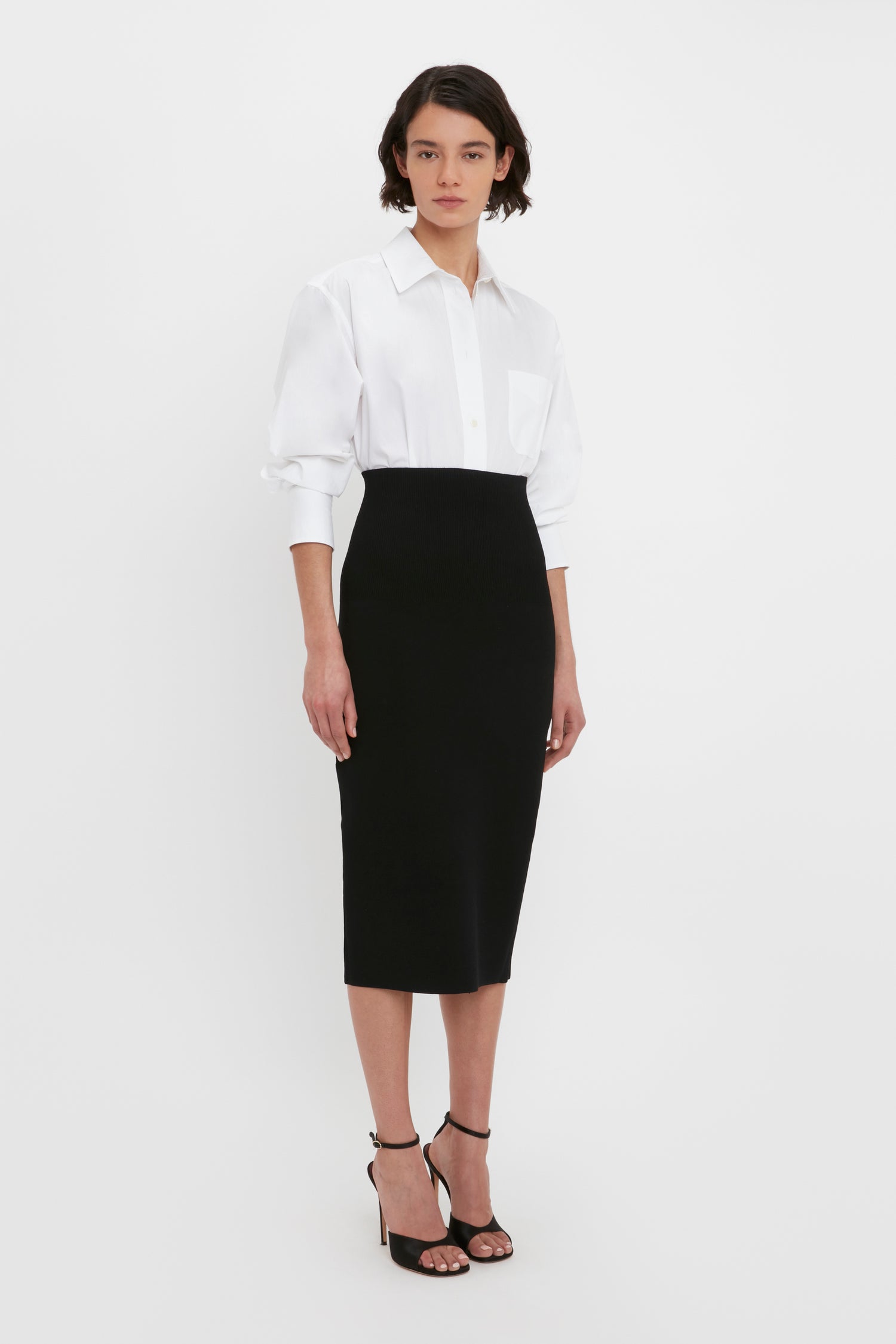 A person stands against a plain background, wearing a white button-up shirt with rolled sleeves and the VB Body Fitted Midi Skirt in Black by Victoria Beckham, paired with black heeled sandals.
