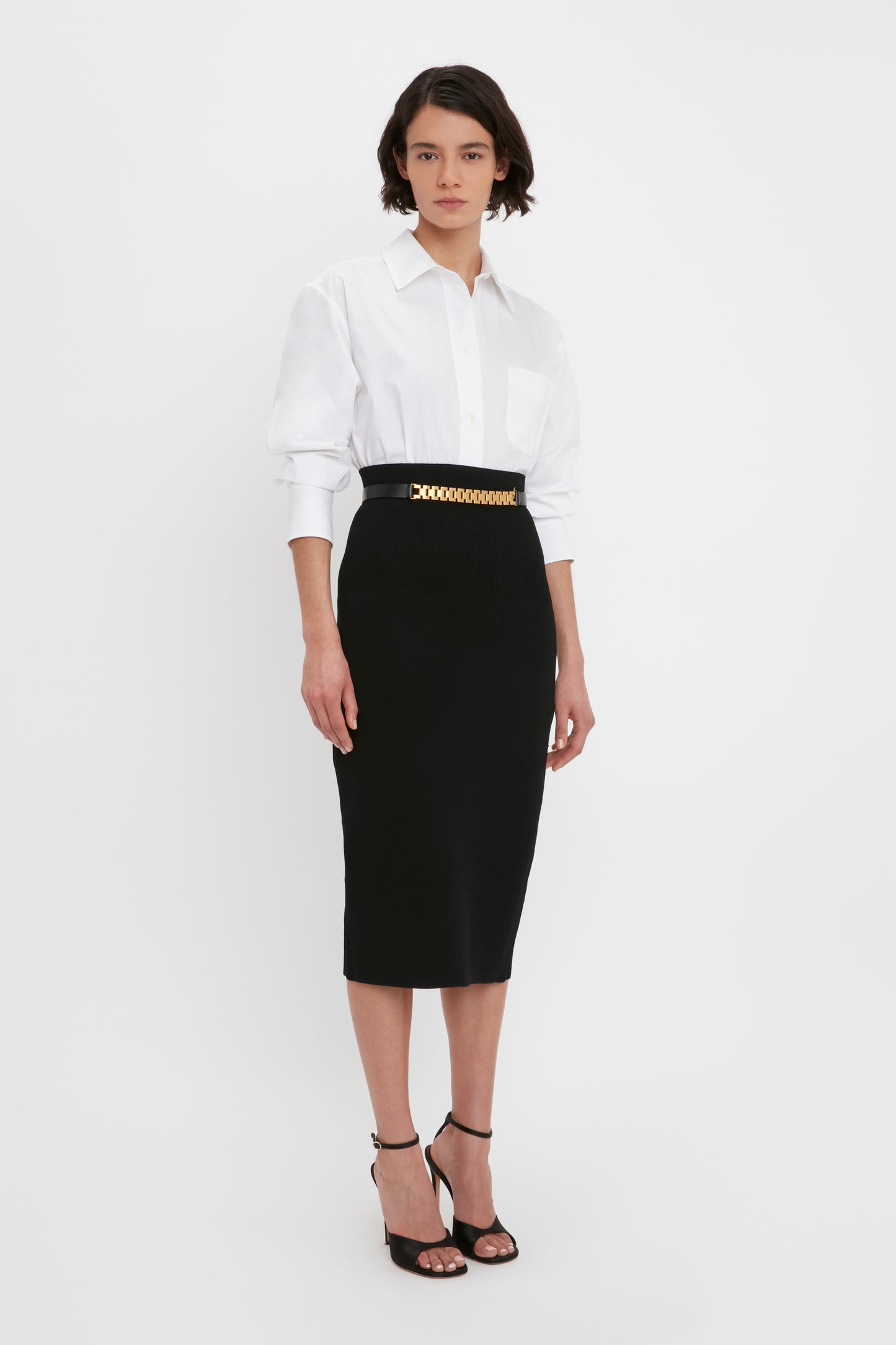 A person is standing against a white background wearing a white button-up shirt, the VB Body Fitted Midi Skirt in Black by Victoria Beckham with a gold belt, and black high-heeled sandals.