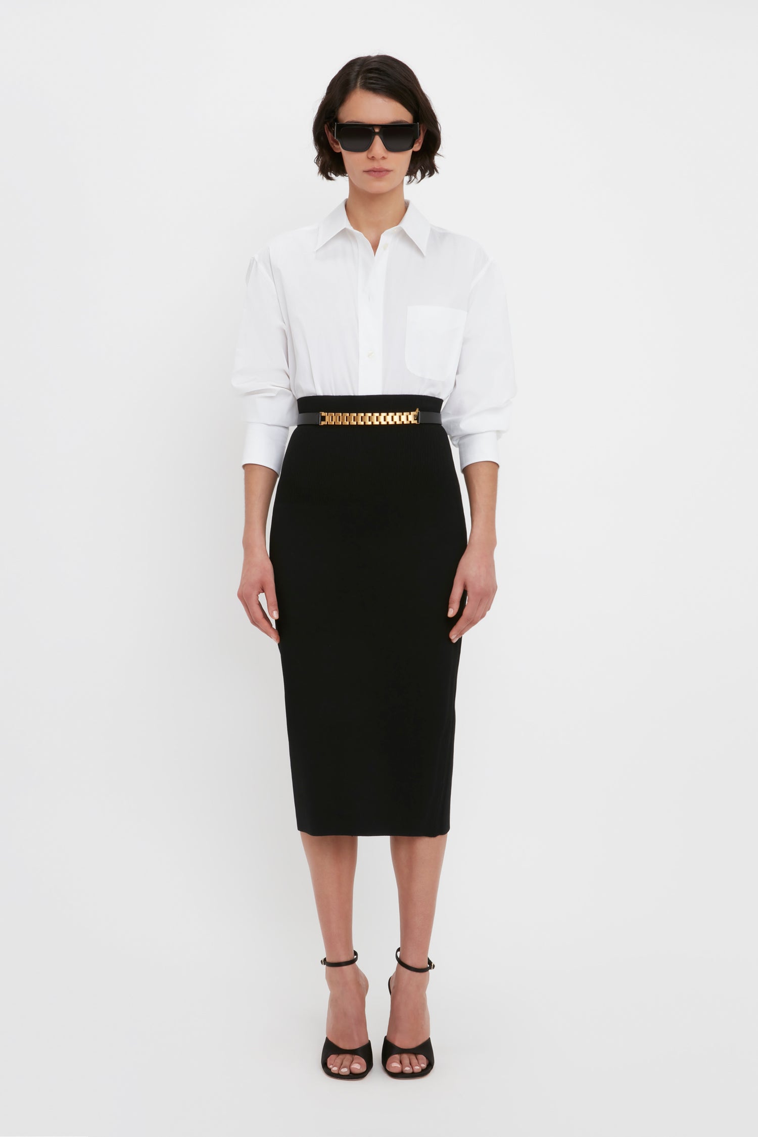 A person stands against a white background wearing sunglasses, a white button-down shirt, a chain belt, black high-heeled sandals, and the VB Body Fitted Midi Skirt in Black from Victoria Beckham.