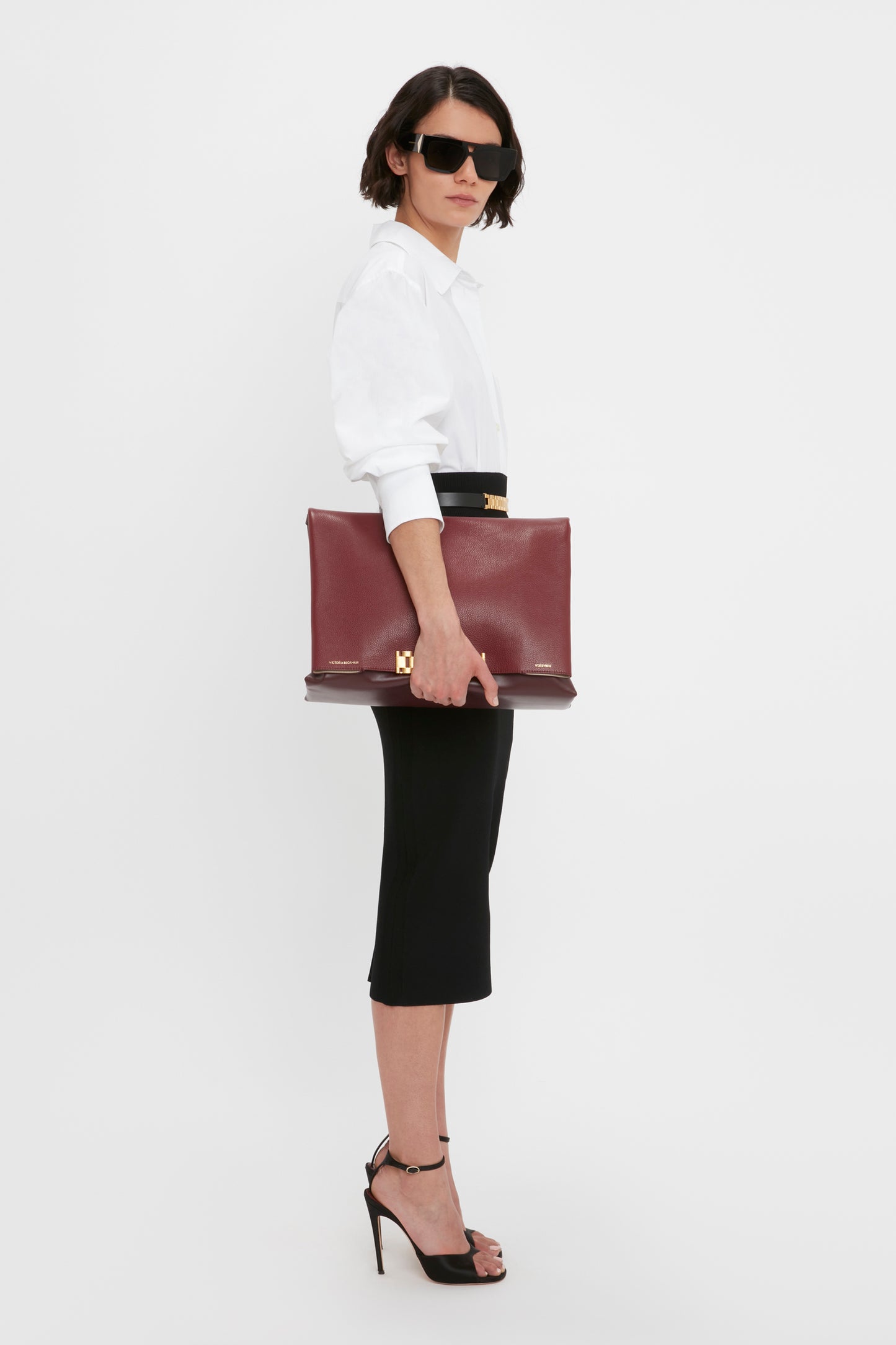 A person in a white shirt and Victoria Beckham's VB Body Fitted Midi Skirt in Black stands with a large red clutch under their arm, wearing black high heels and dark sunglasses. The background is plain white.