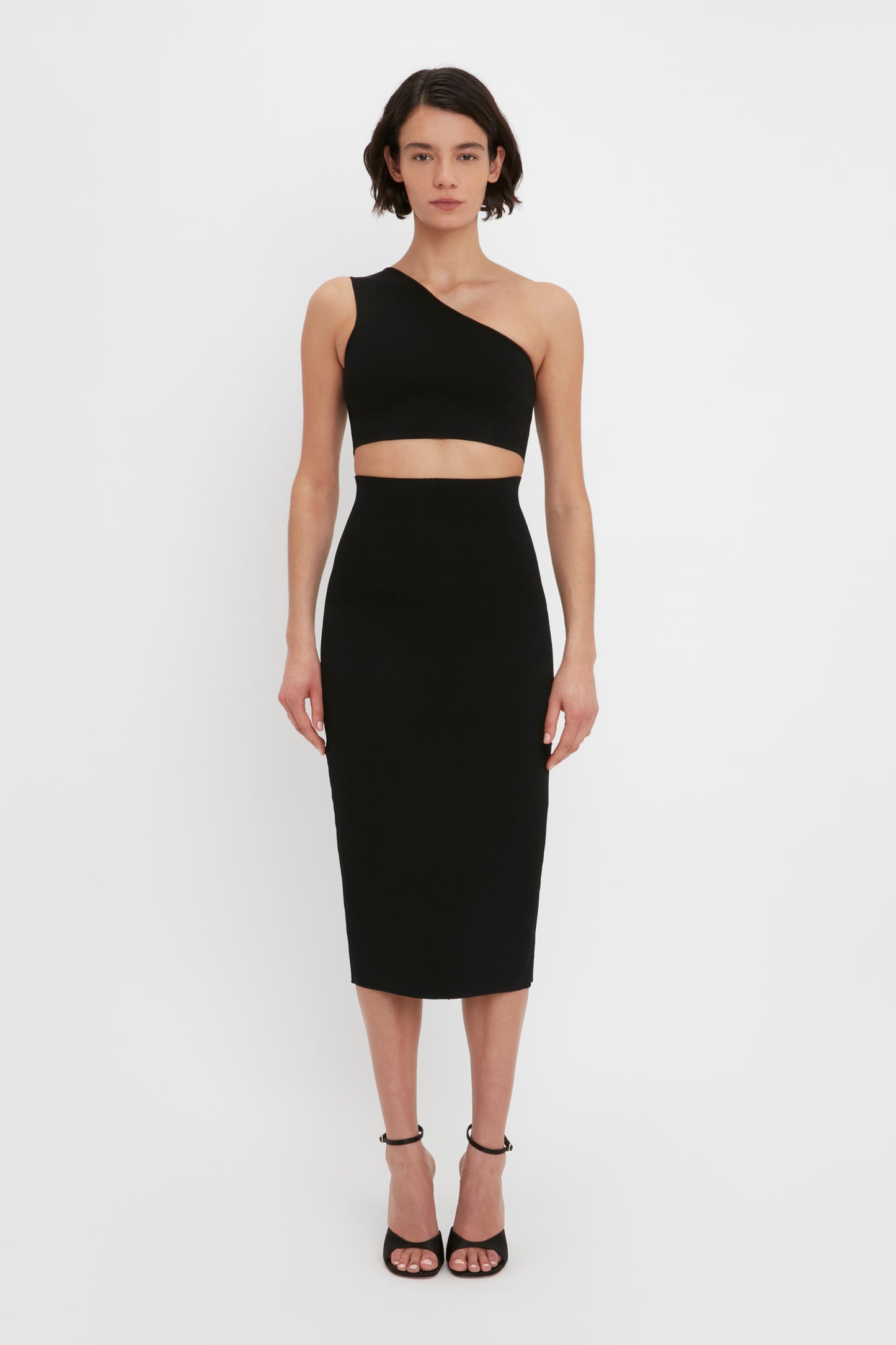 A person stands against a white background, wearing the VB Body Fitted Midi Skirt in Black by Victoria Beckham paired with black high-heeled sandals.