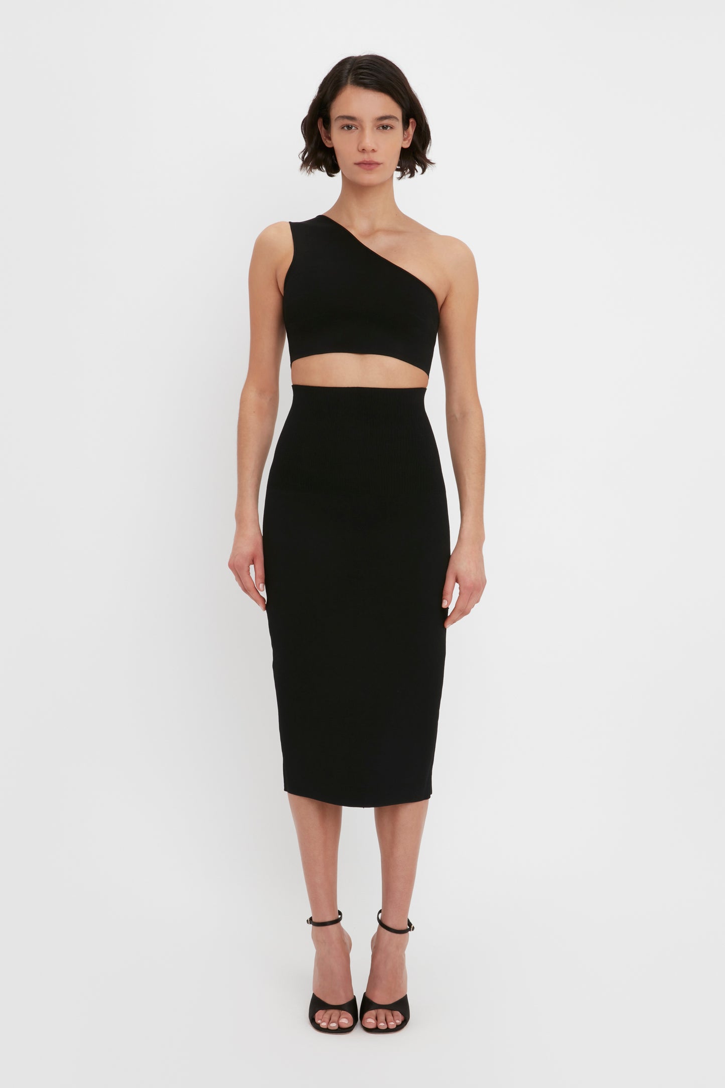 Person wearing a VB Body One Shoulder Crop Top in Black by Victoria Beckham and a matching high-waisted midi skirt with open-toe heels, standing against a plain white background.