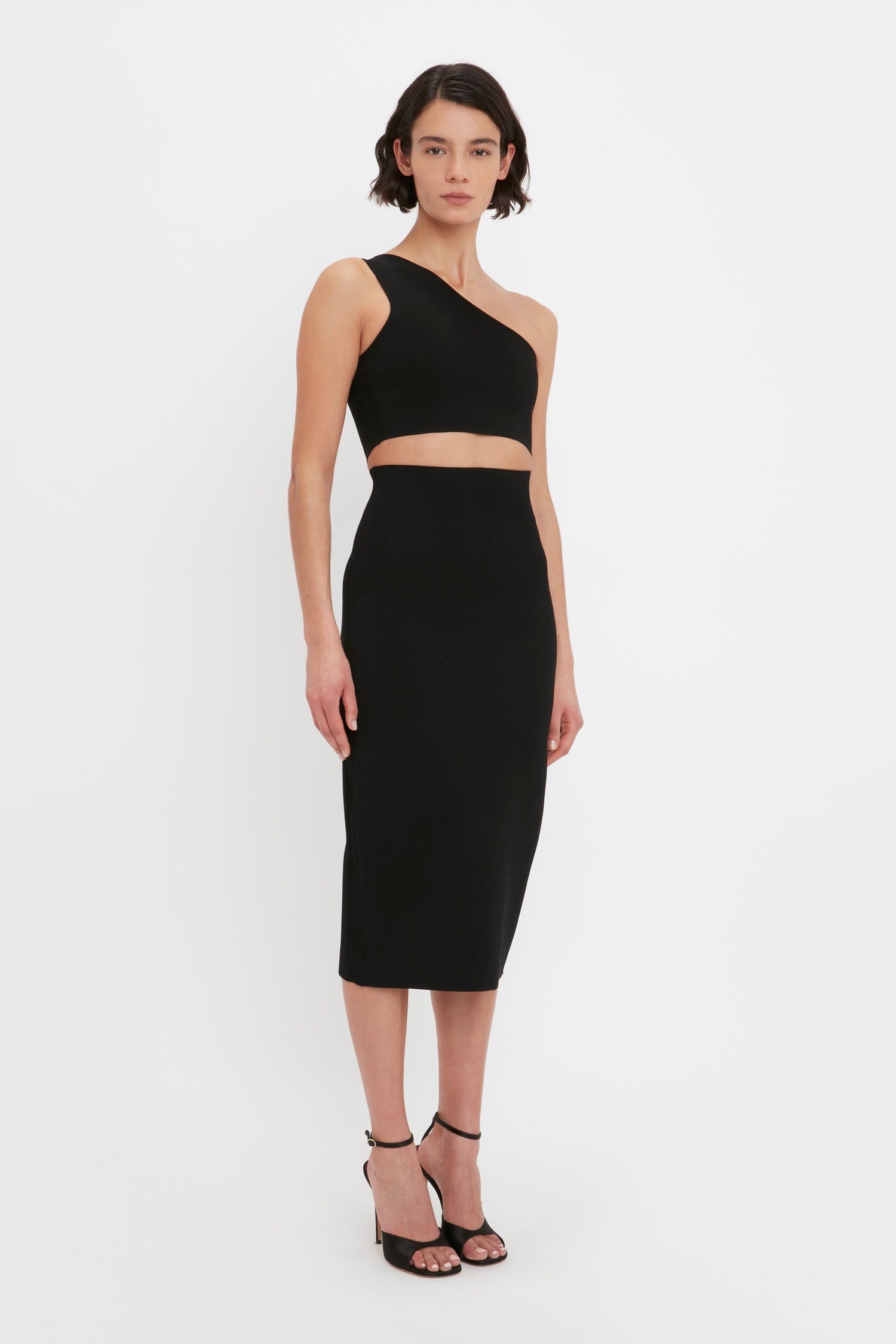A person with short dark hair, wearing a black one-shoulder VB Body Crop Top by Victoria Beckham and black high-heeled sandals, standing against a white background.