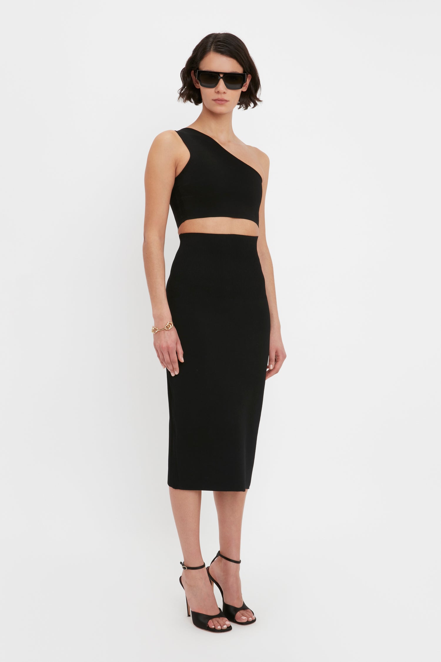 Person wearing a one-shoulder black crop top and the Victoria Beckham VB Body Fitted Midi Skirt in Black, accessorized with black heels, sunglasses, and a bracelet, standing against a white background.