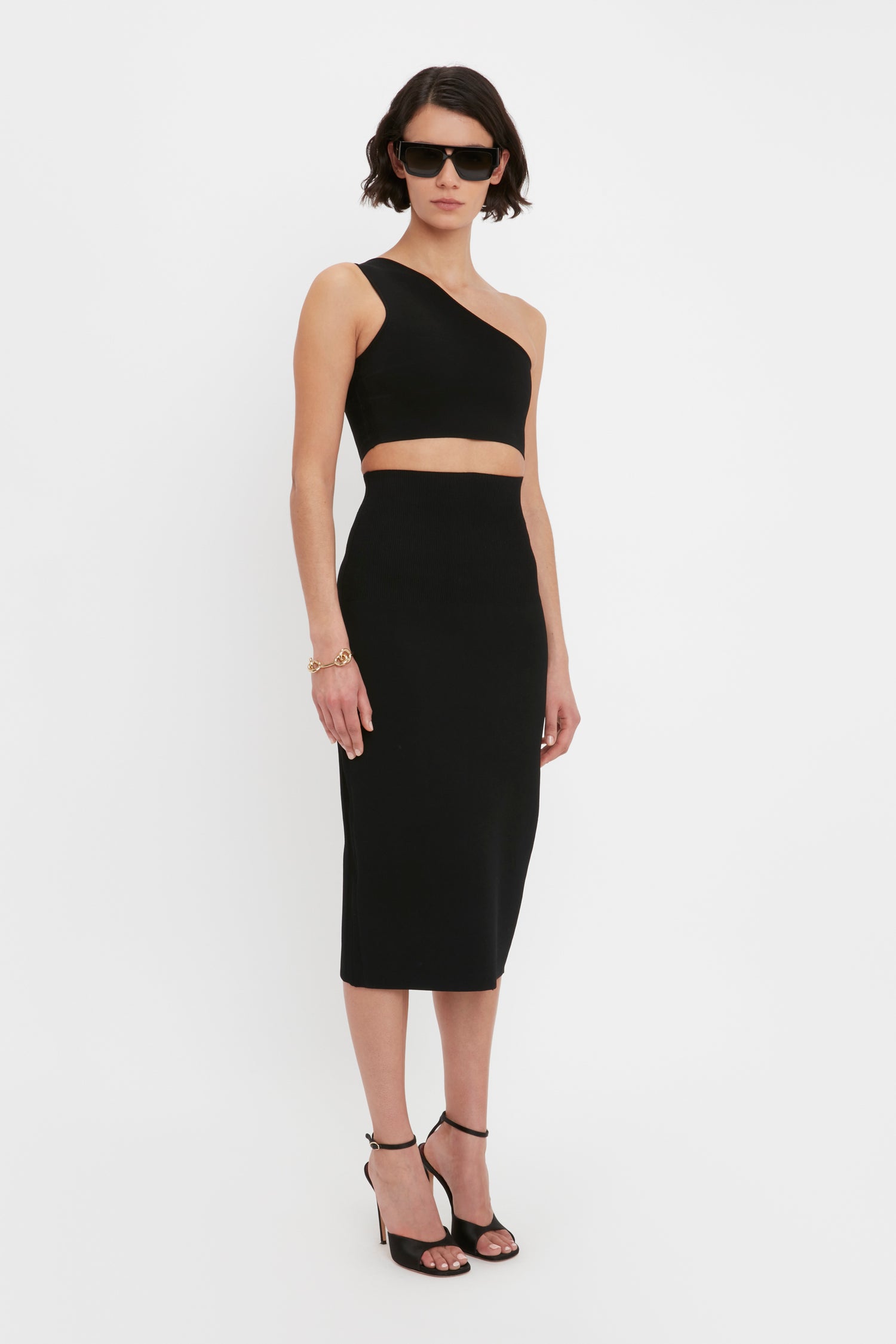 A person with short dark hair wearing sunglasses, the VB Body One Shoulder Crop Top by Victoria Beckham in black, and a matching black midi skirt with high heels, standing against a plain white background.