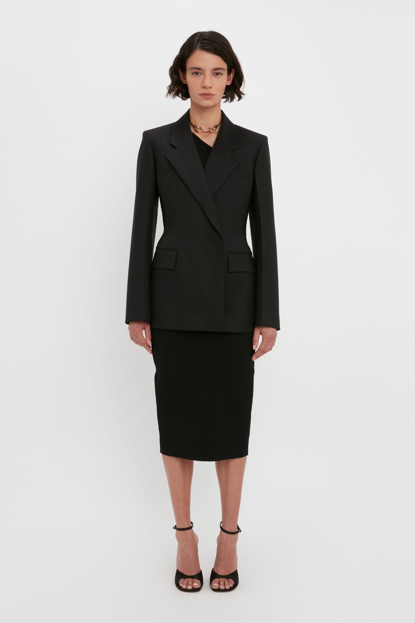 A person with short dark hair stands against a white background wearing the VB Body One Shoulder Crop Top in Black from Victoria Beckham, a matching skirt, open-toe heels, and a gold chain necklace.