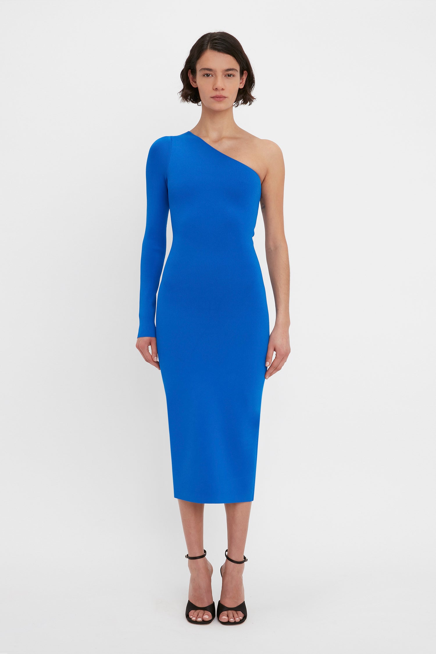 A woman stands in a Victoria Beckham VB Body One Shoulder Midi Dress in Cobalt and black high-heeled sandals against a white background.