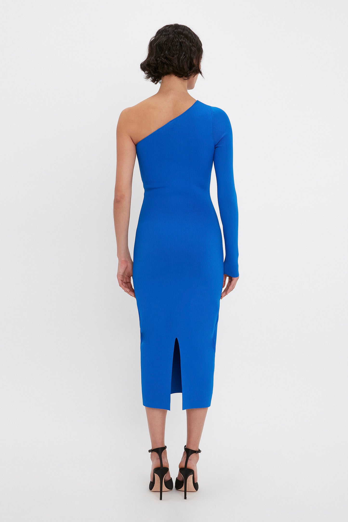 A person is wearing the Victoria Beckham VB Body One Shoulder Midi Dress in Cobalt, featuring a single long sleeve and a slit at the hem, viewed from the back.